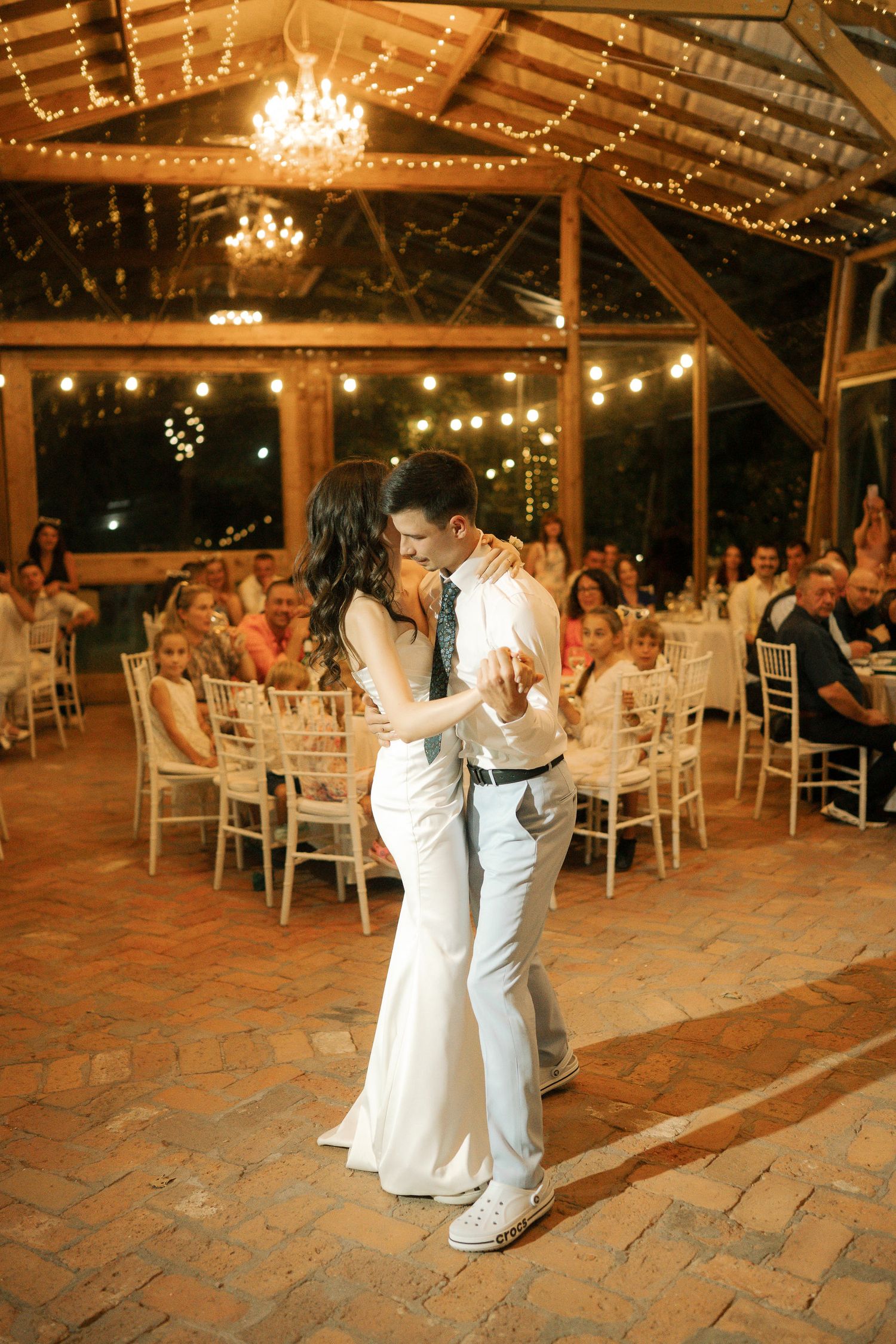 A couple shares their first dance at a rustic barn wedding reception with string lights and chandeliers overhead.