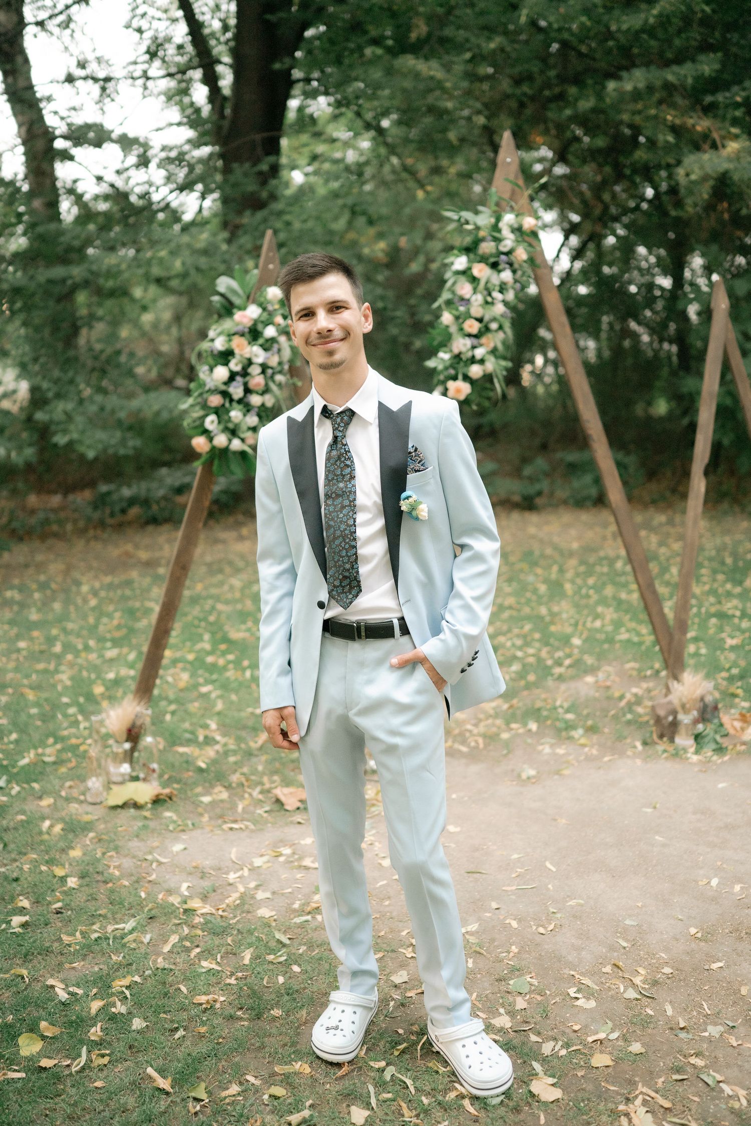 A young boy in a light gray suit and sneakers poses playfully in an outdoor setting with autumn leaves on the ground.