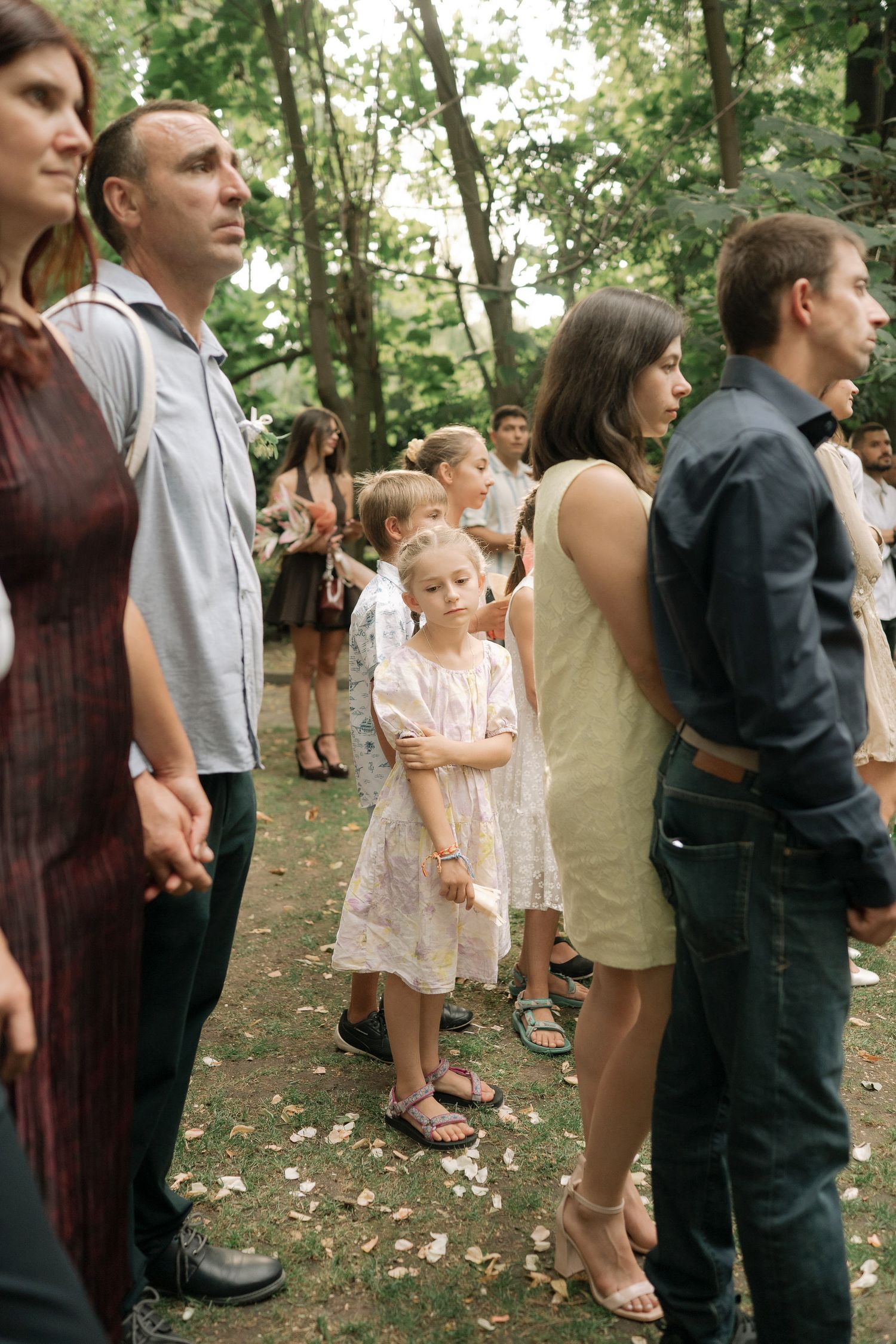 People gather outdoors at a casual event in a wooded area wearing mix of formal and casual attire.
