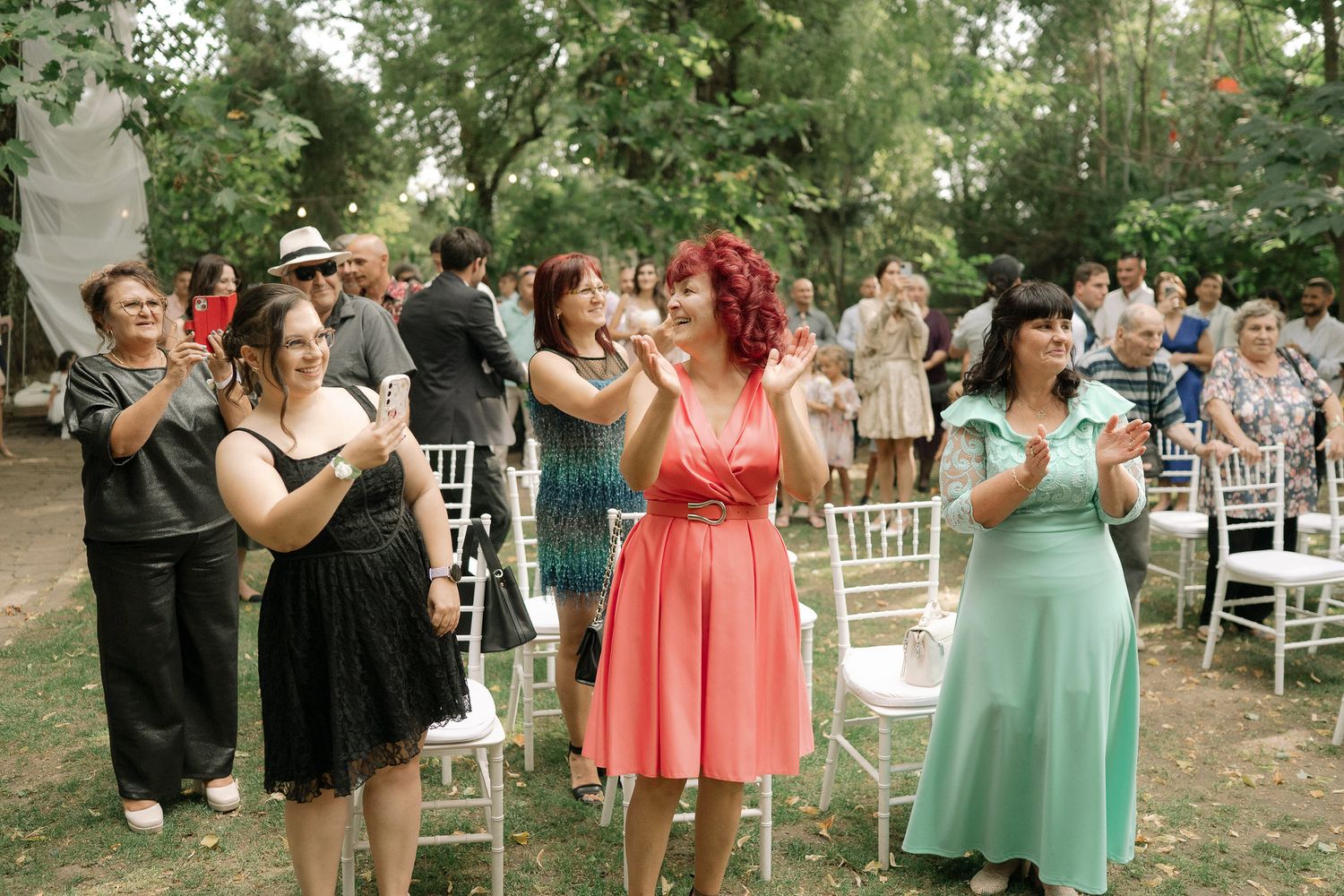 Wedding guests dance together at an outdoor reception with white chairs and trees in the background.
