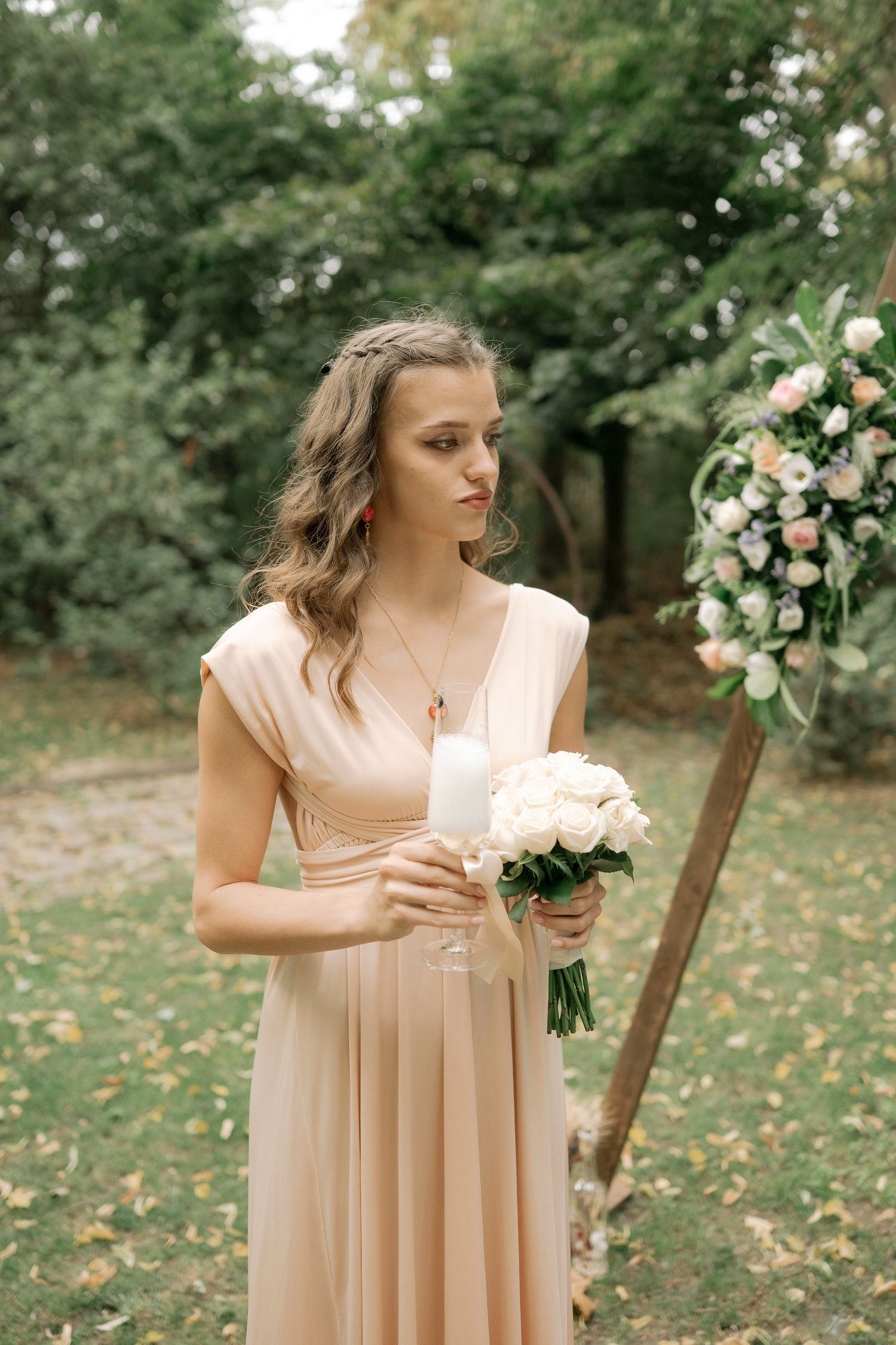 A bridesmaid in a blush pink dress holds a white rose bouquet in an outdoor garden setting with floral decorations.