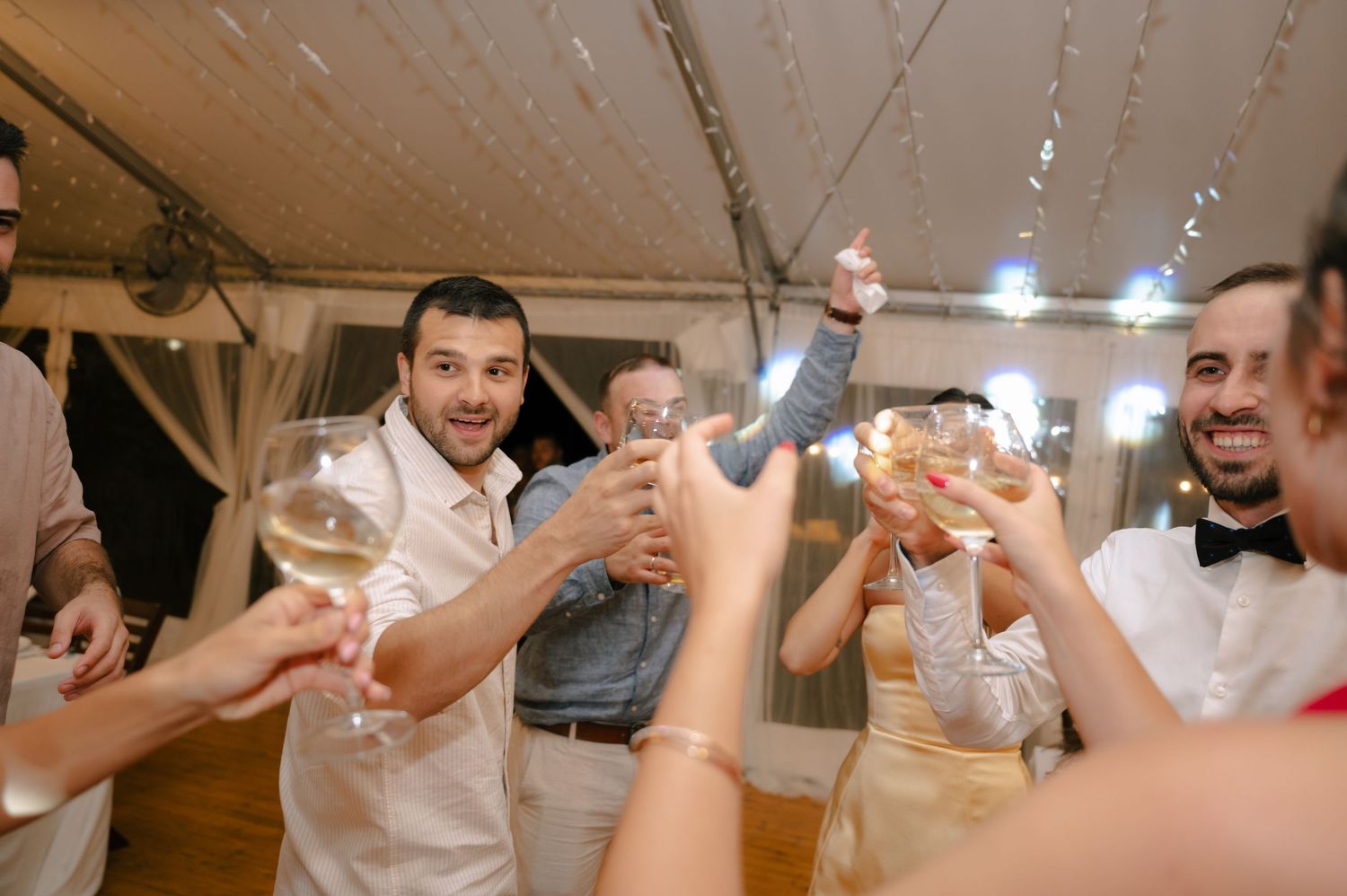 Wedding guests raise their glasses in a celebratory toast under a tented reception space.