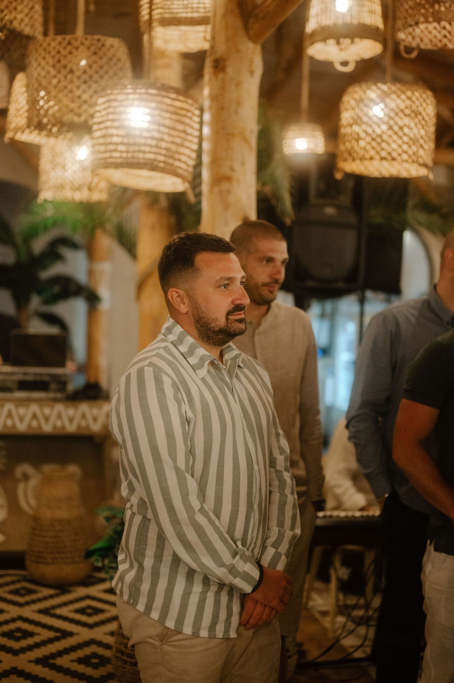 A person in a striped shirt stands in a warmly lit restaurant interior with crystal chandeliers overhead.