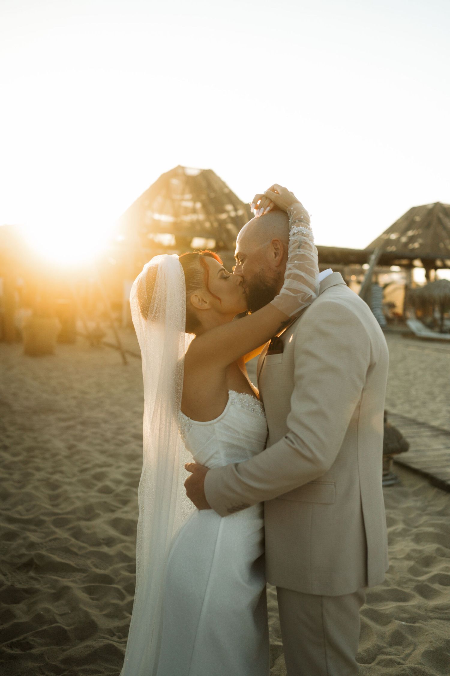 A couple shares a romantic kiss on a sandy beach at sunset with warm golden light illuminating their silhouettes.