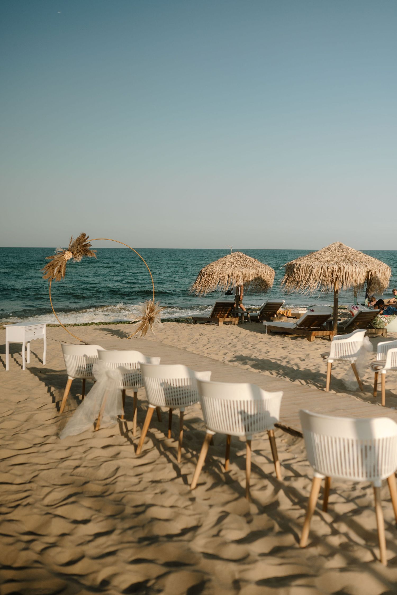 Beach lounge setup with white chairs and thatched umbrellas along a sandy shoreline at sunset.