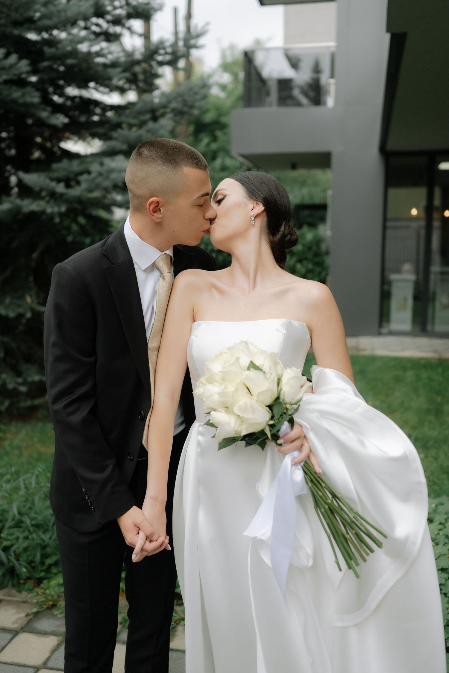 A romantic wedding couple shares a tender kiss outdoors, with the bride holding a bouquet of white roses.