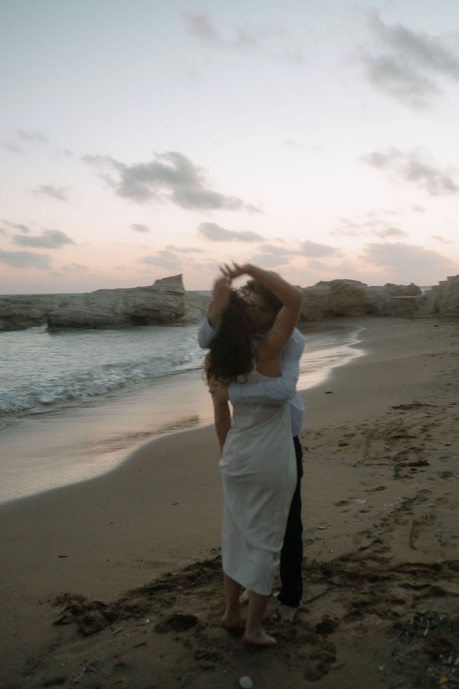 A couple embraces on a sandy beach at sunset, with rocky cliffs and gentle waves in the background.