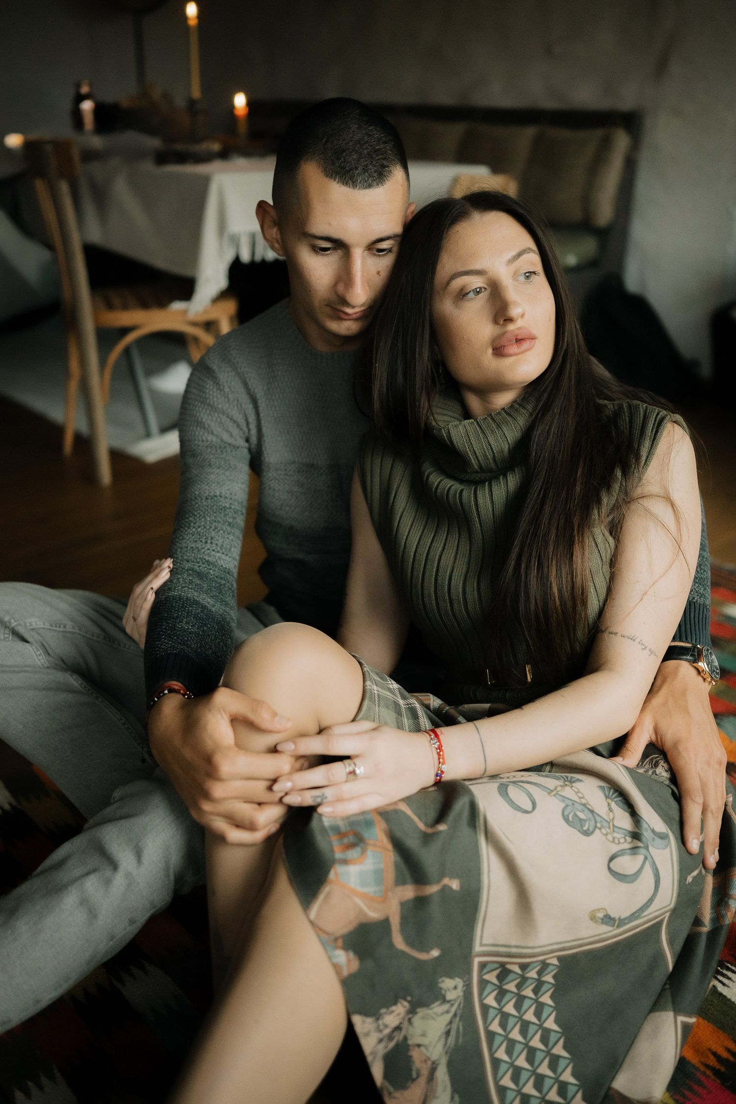A couple sits close together on a couch showing their arm tattoos in a dimly lit room.