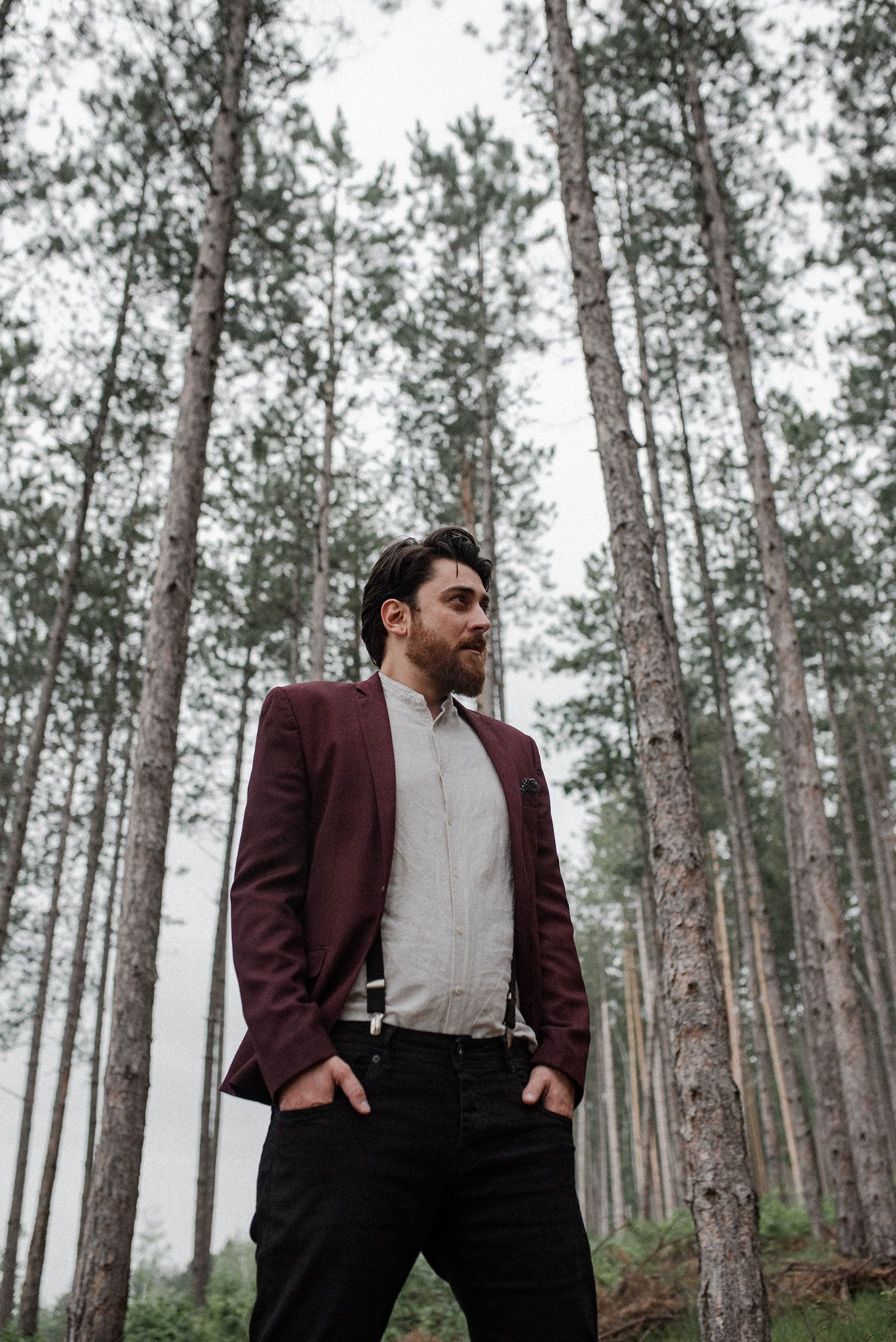 A person in a burgundy blazer and suspenders poses thoughtfully in a misty pine forest setting.
