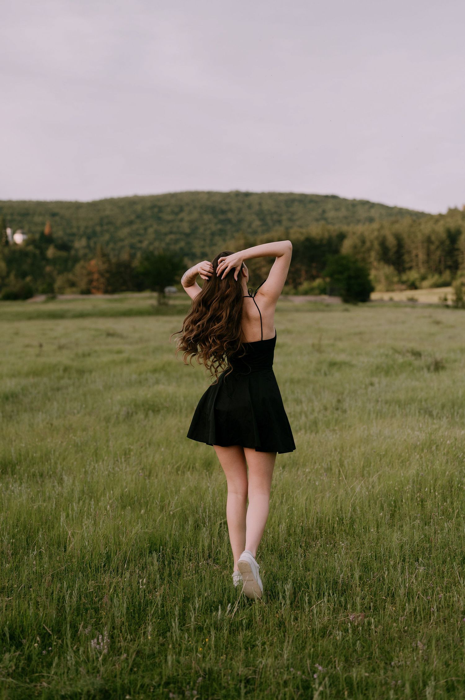 A figure in a black dress dances and twirls through a serene meadow against rolling green hills on a cloudy day.
