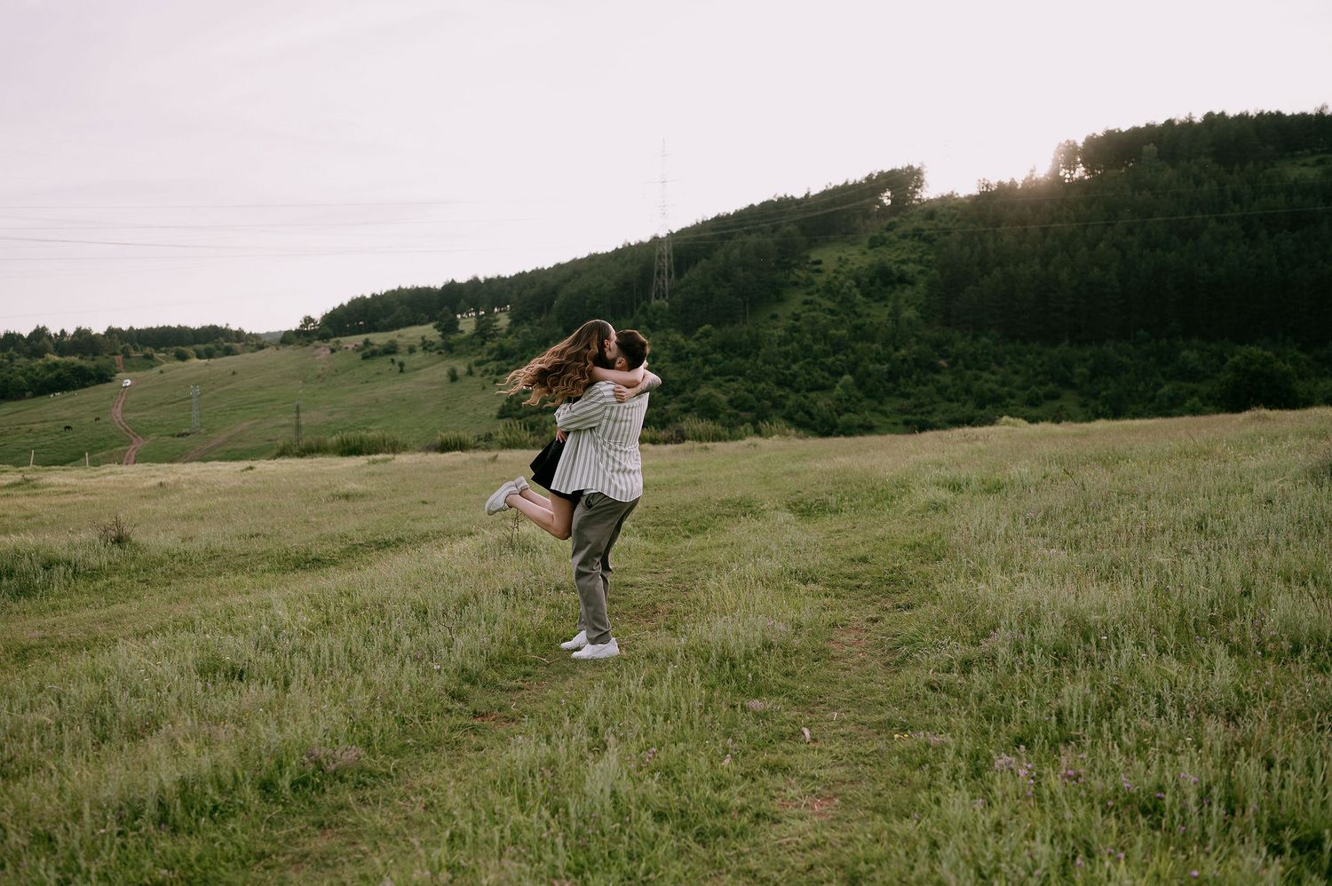 A person dances joyfully in a lush green meadow at sunset with rolling hills in the background.
