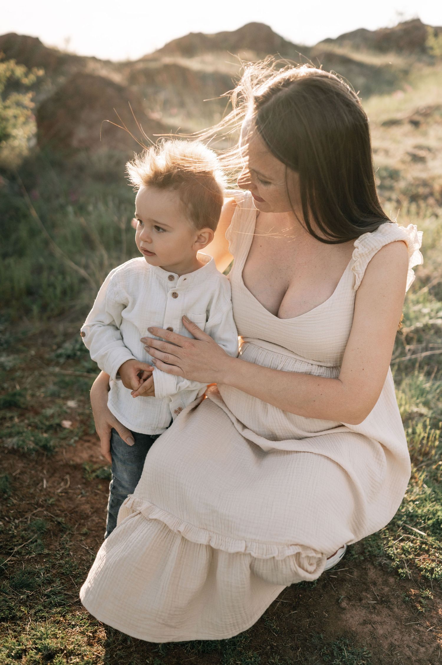 A mother and young child share tender moments together outdoors in warm sunlight wearing cream colored clothing.