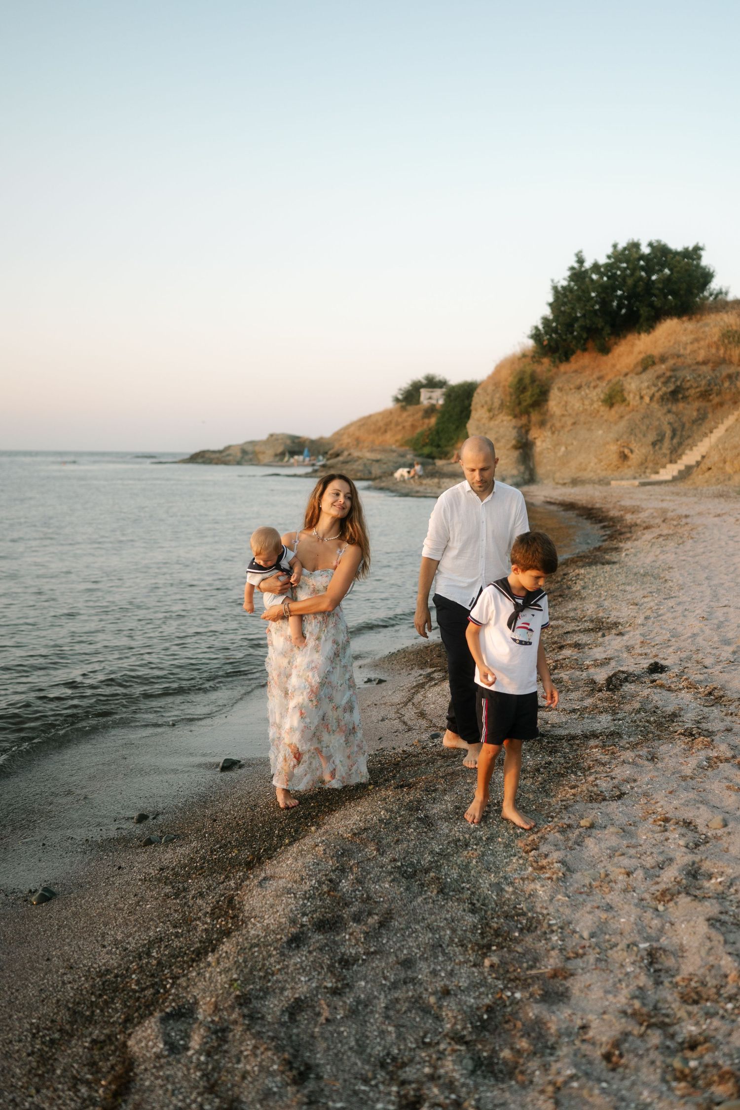 People walking and playing along a scenic beach shoreline with rocky cliffs at sunset.