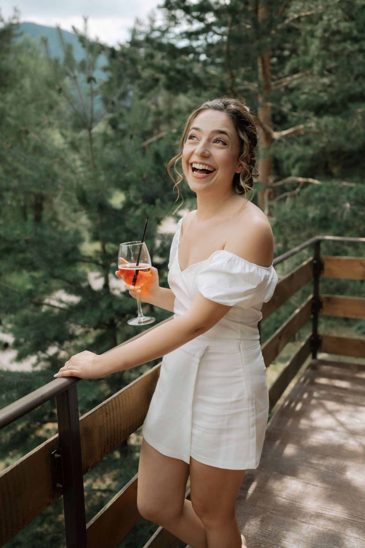 A woman in a white summer outfit sits and smiles on a wooden deck surrounded by pine trees in a mountain setting.