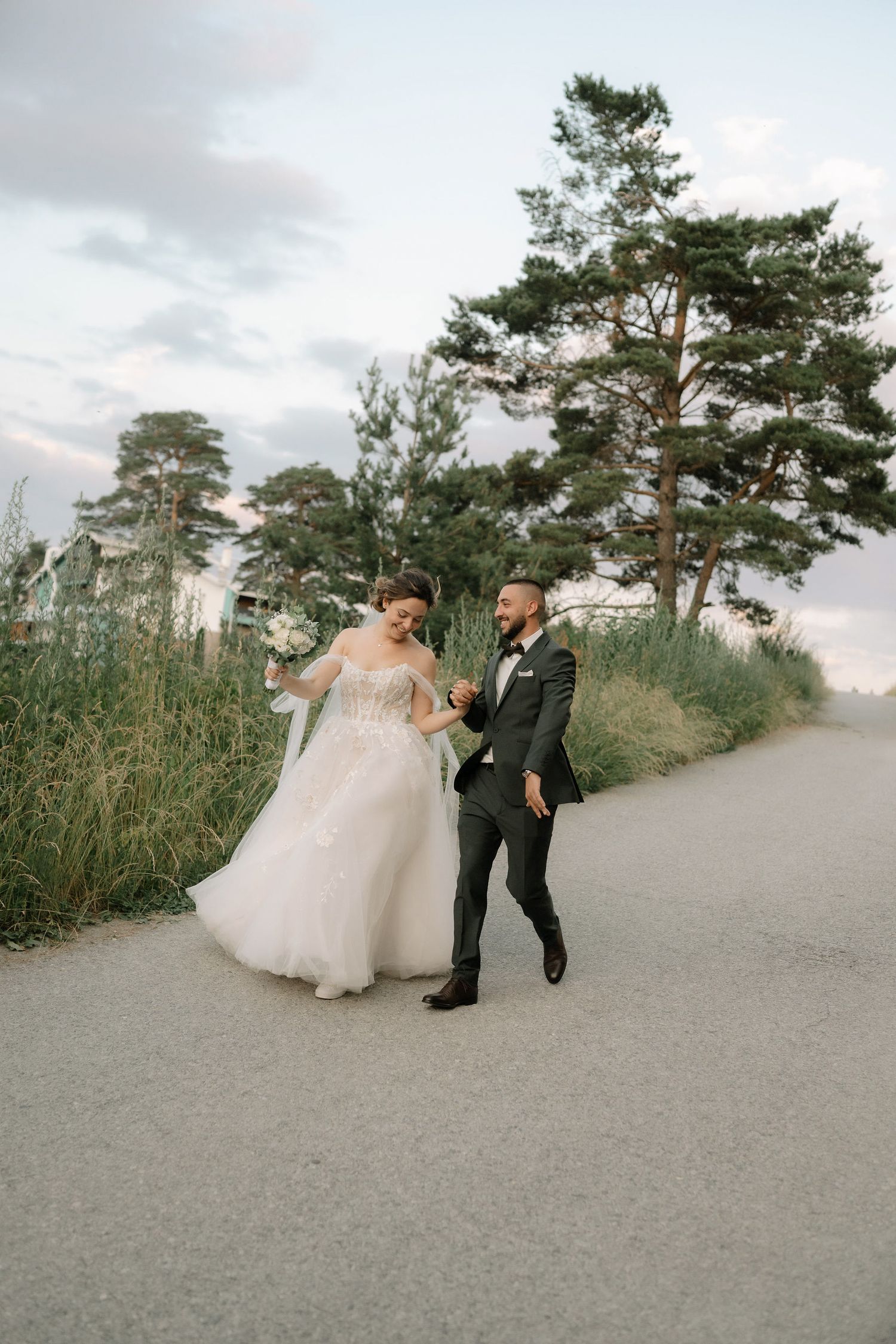 A couple in wedding attire share a playful moment on a path lined with tall pine trees on an overcast day.