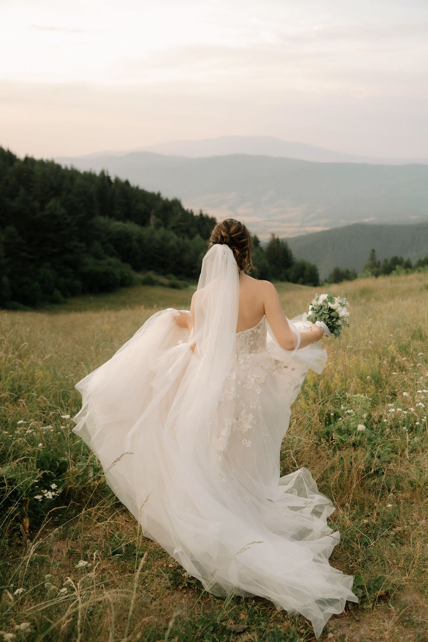 A bride in a flowing white dress holds a bouquet while standing on a hillside overlooking misty mountain ranges.