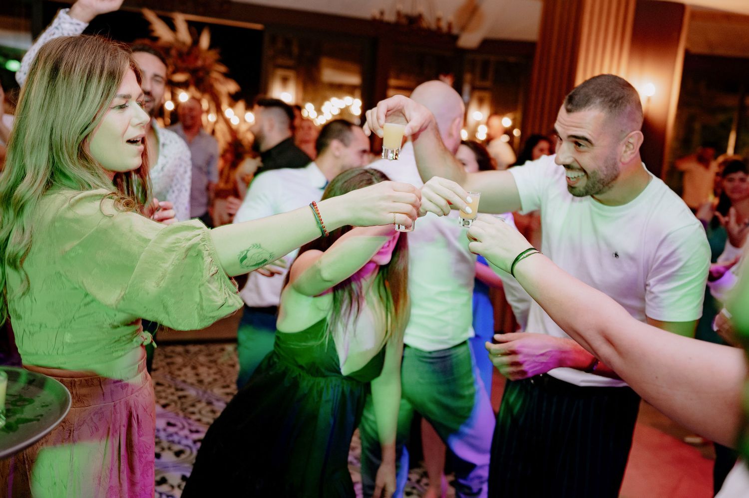 People dancing and celebrating at a party with green lighting in a crowded indoor venue.