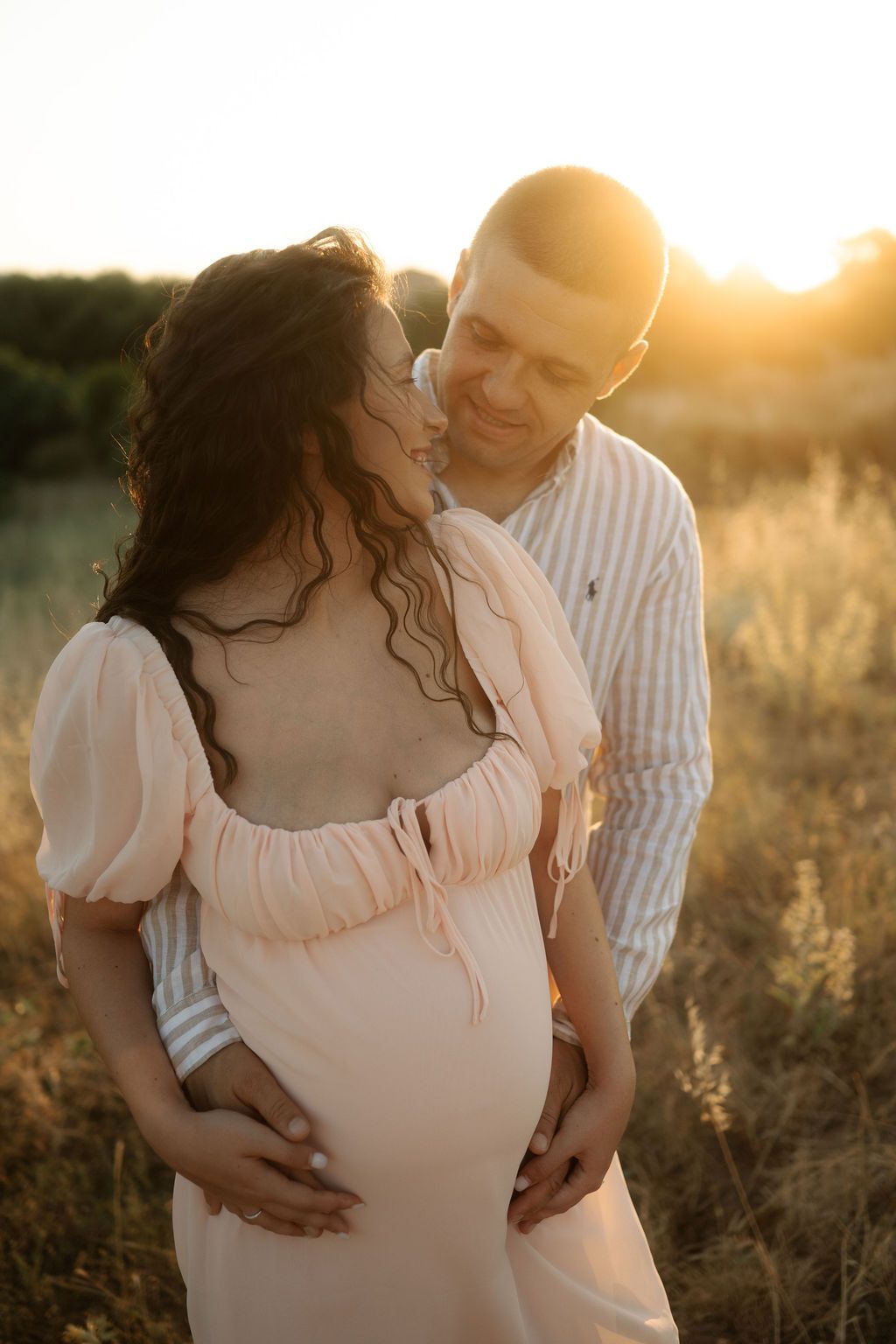 Expectant couple shares loving embrace during sunset maternity session in rustic field setting.
