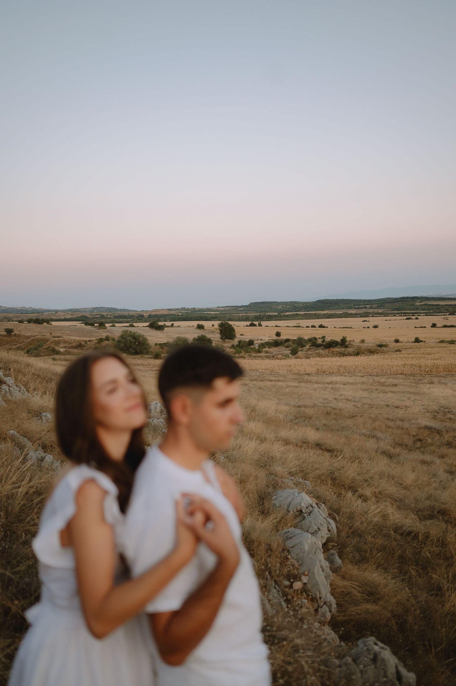A couple embraces in a golden field at sunset with a vast prairie landscape stretching to the horizon.