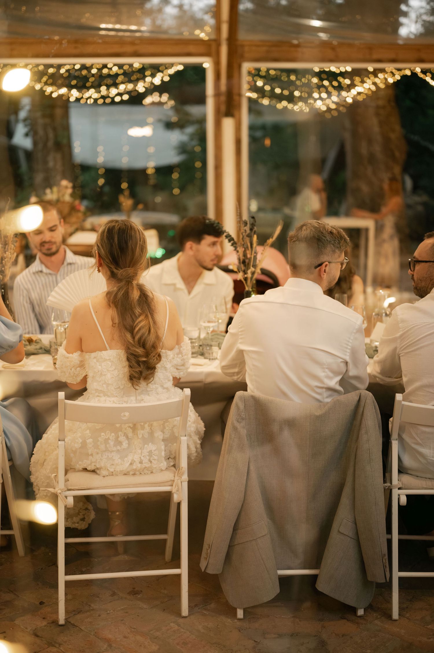 Wedding guests enjoy an intimate dinner reception under twinkling lights in a glass-enclosed venue.