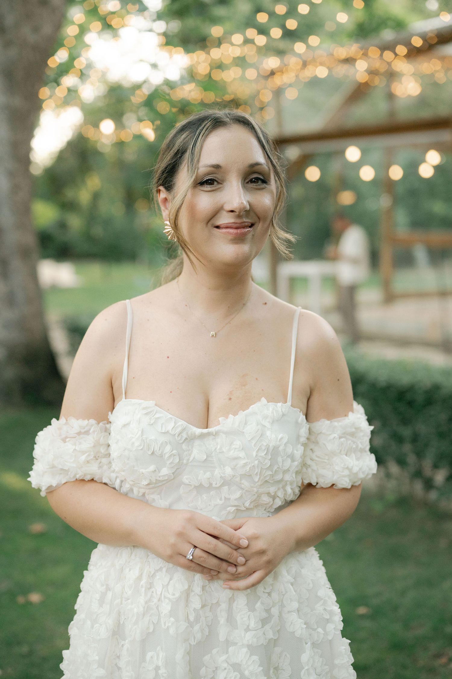 A bride in a white off-shoulder floral wedding dress poses outdoors with string lights and greenery in the background.