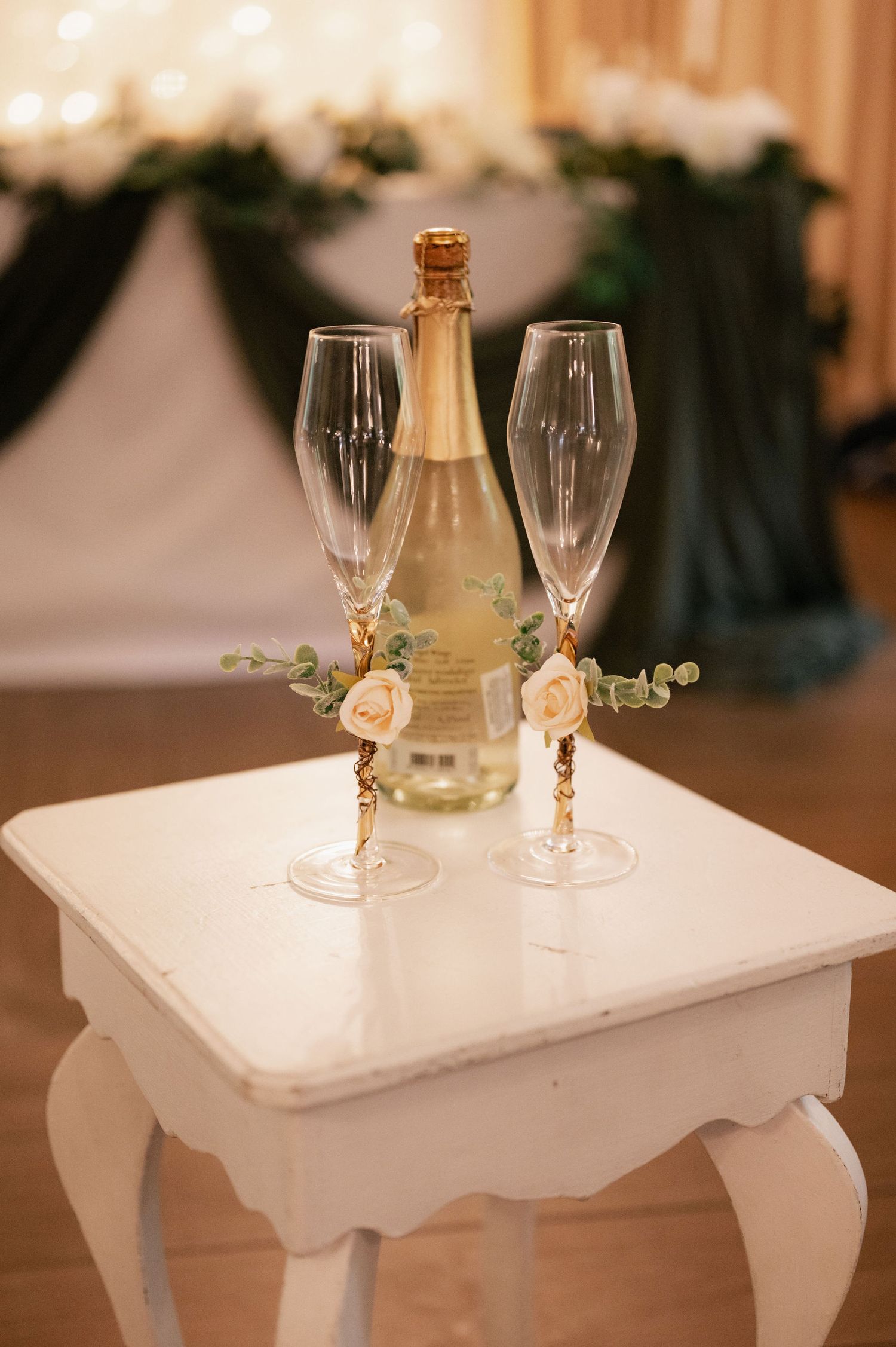White decorative table with champagne glasses and floral accents set against a warm-toned wedding reception backdrop.