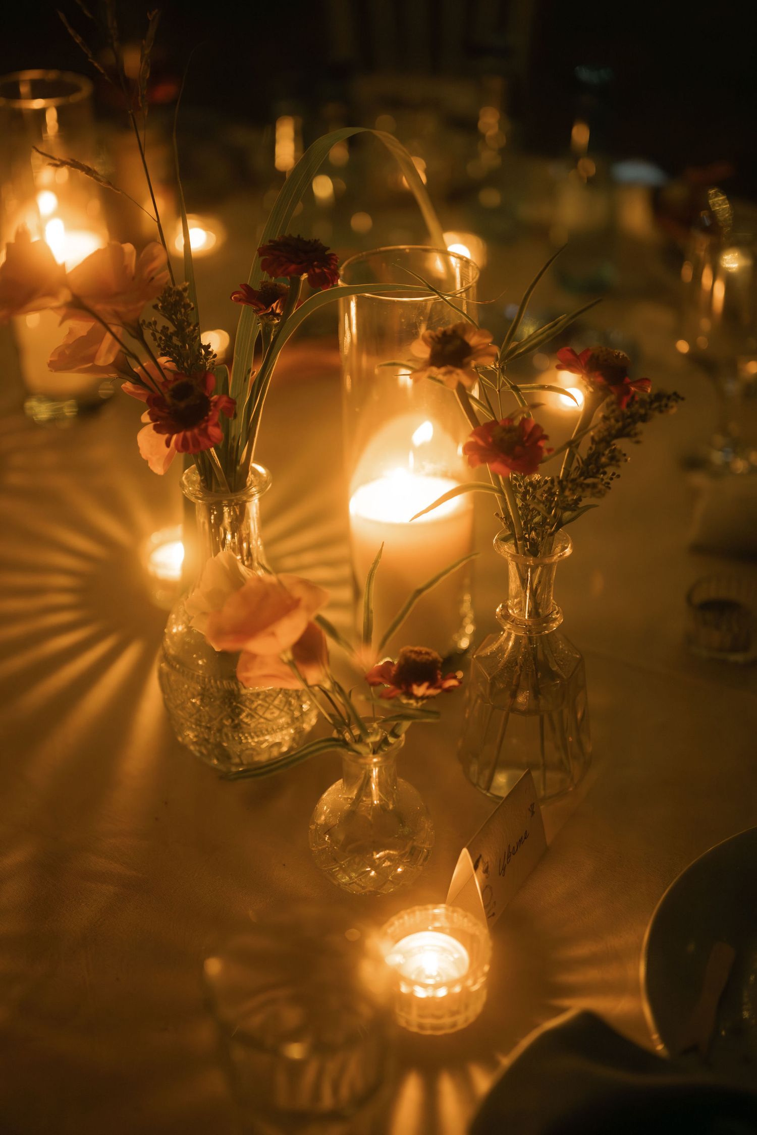 Glowing tea lights cast radiant patterns on a dark surface with delicate flower arrangements in small glass vases.