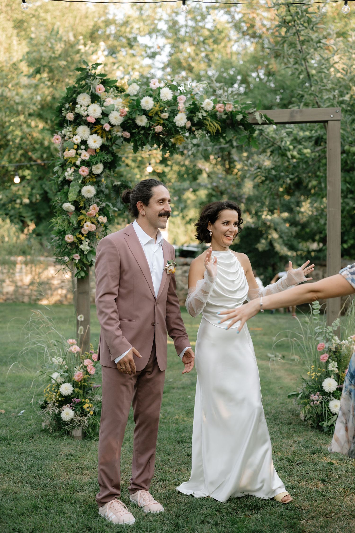A couple in wedding attire dances playfully in a garden setting decorated with floral arrangements and greenery.