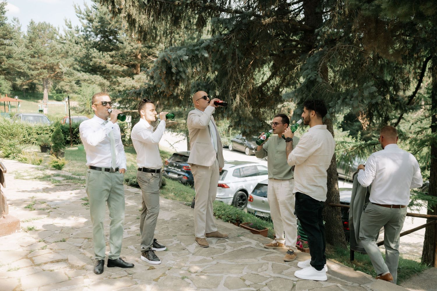 Wedding party members in light gray and sage green attire gather outdoors under trees for group photos.