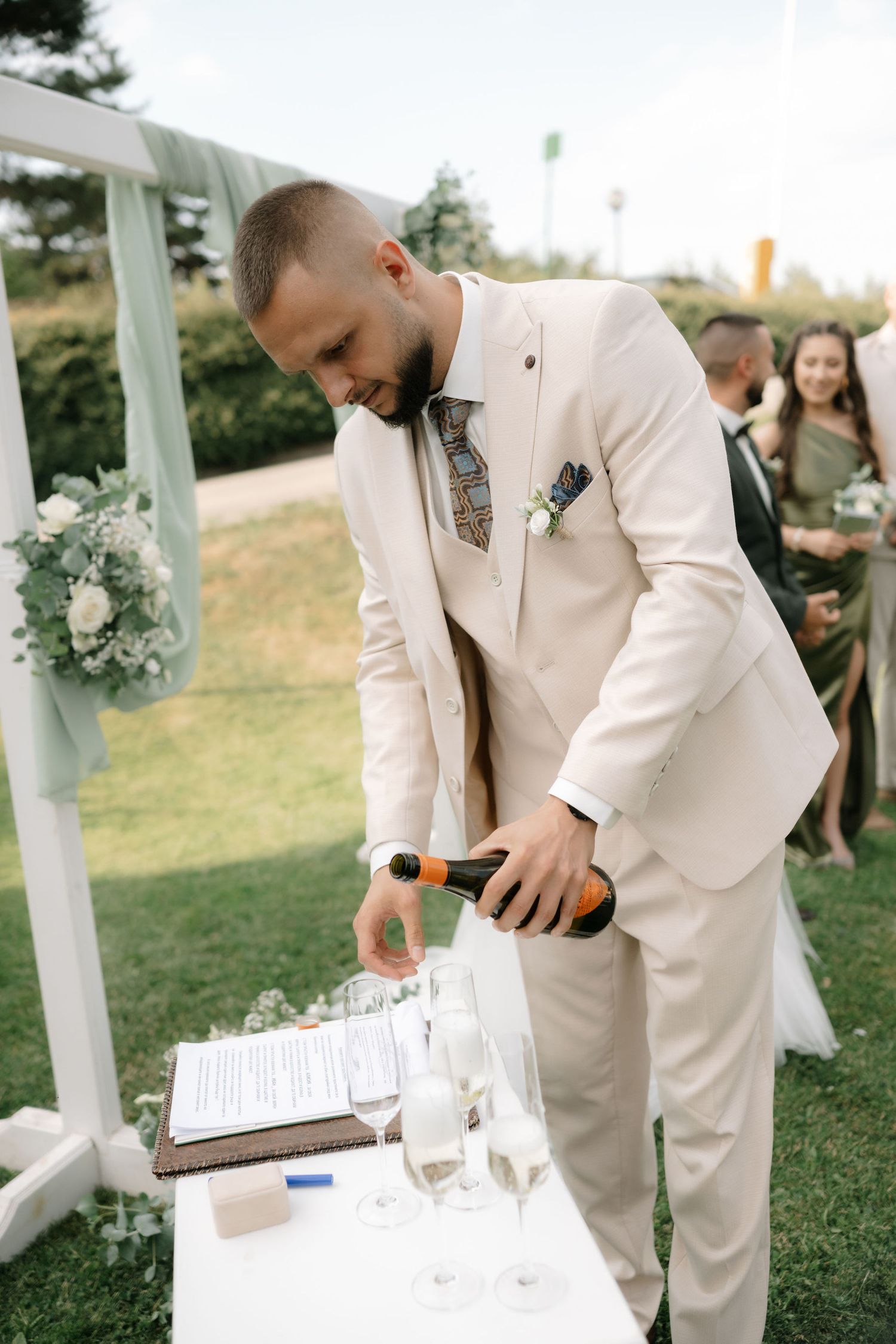 Groomsmen in light colored suits prepare a wedding reception table with glasses and settings outdoors.