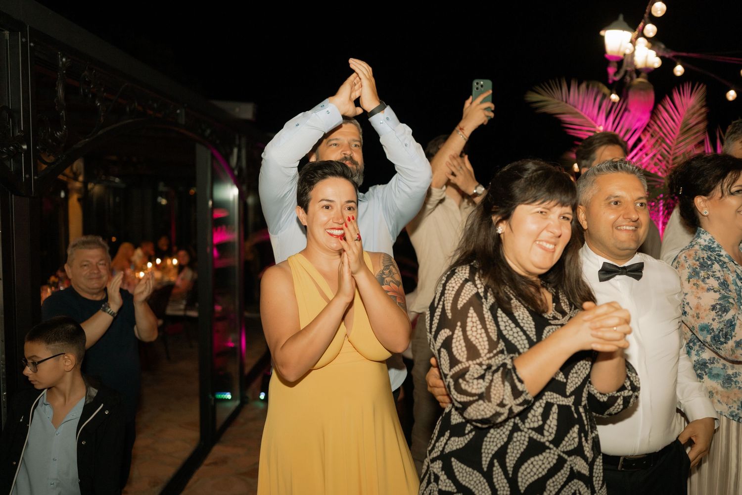 Party guests dance and celebrate with raised arms on a lively dance floor with colorful lighting in the background.