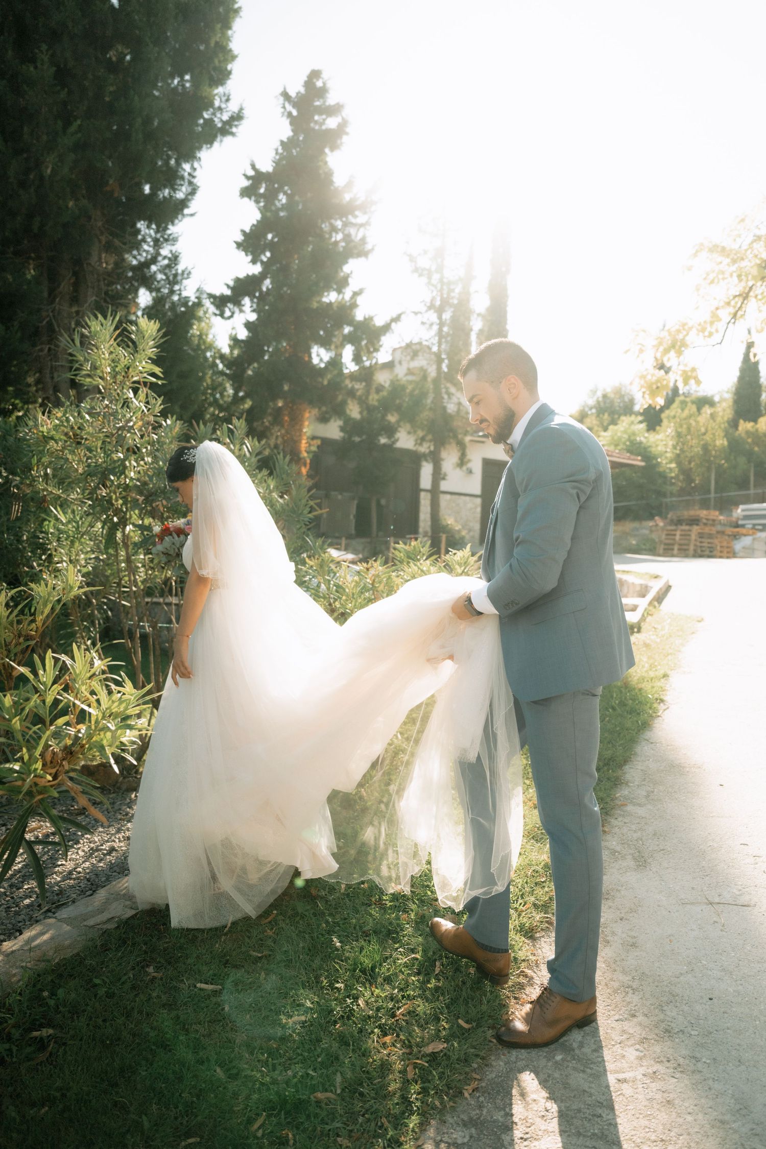 A couple in wedding attire shares a romantic moment on a sunlit garden path as the bride's dress flows in the breeze.
