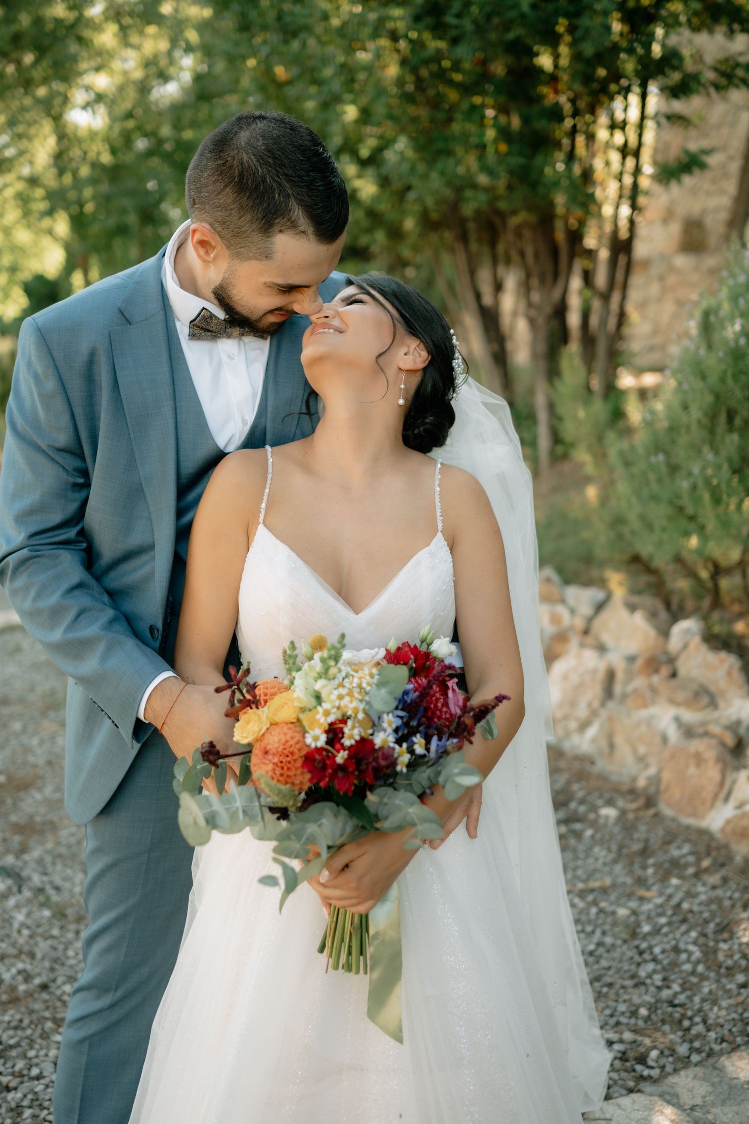 A romantic wedding couple shares tender moments in a wooded outdoor setting with autumn-colored floral bouquet details.