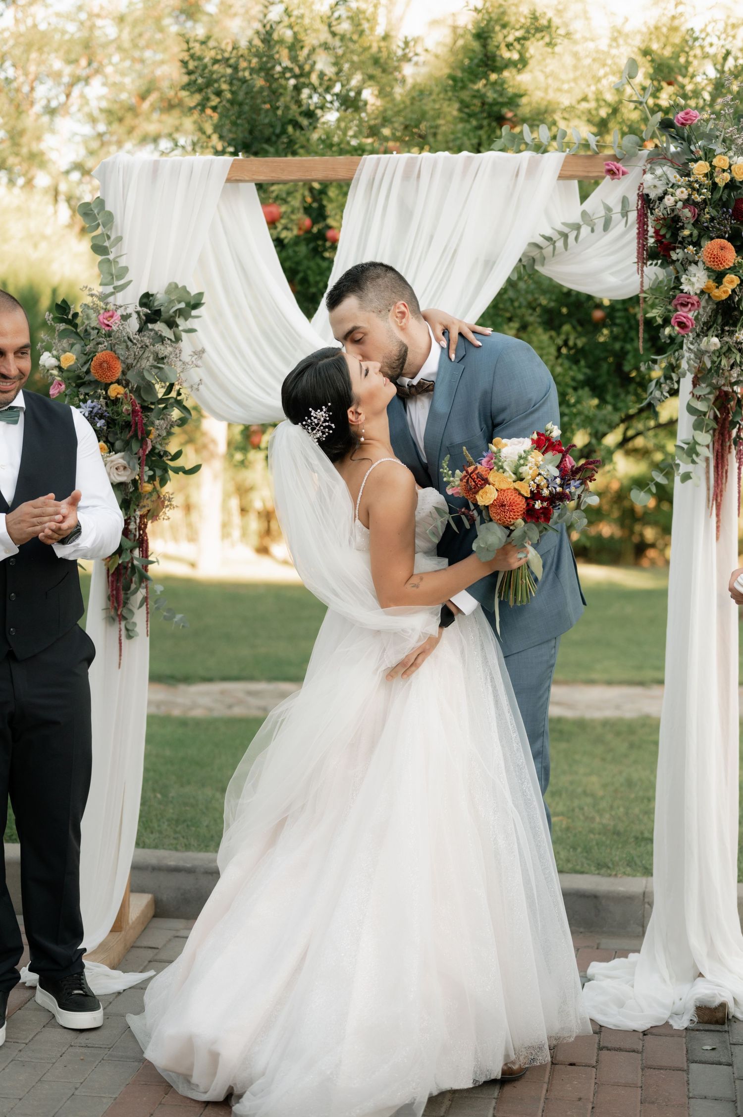 A romantic outdoor wedding ceremony under a white draped arch decorated with vibrant orange and pink florals.