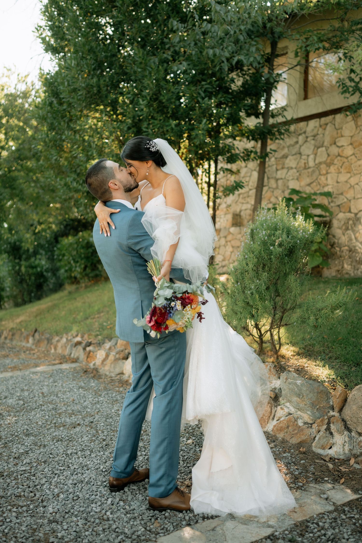 A romantic wedding couple embraces and dances together on a gravel path during golden hour with stone walls in the background.