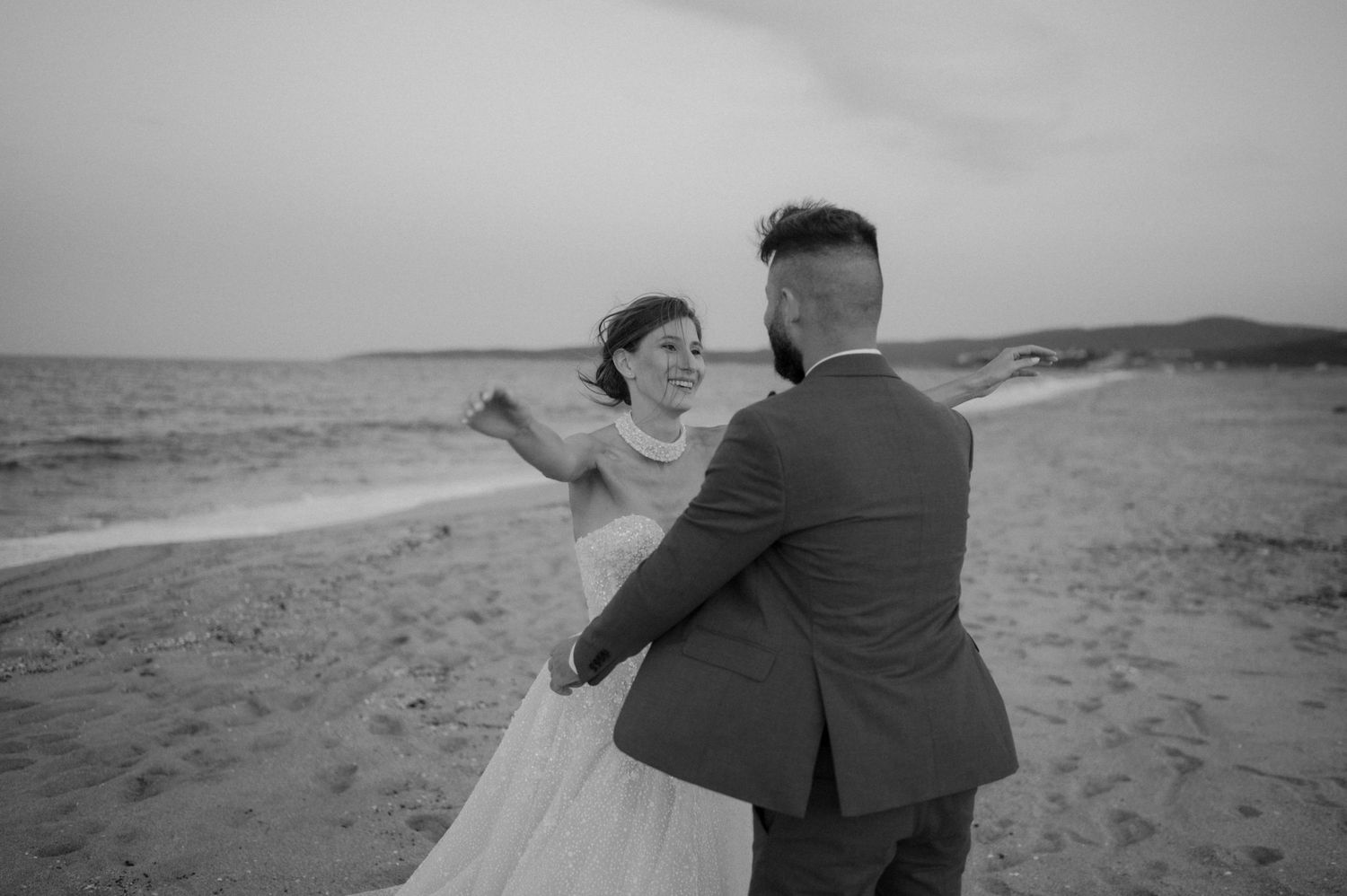 Black and white photo of a bride and groom dancing romantically on a sandy beach by the ocean.