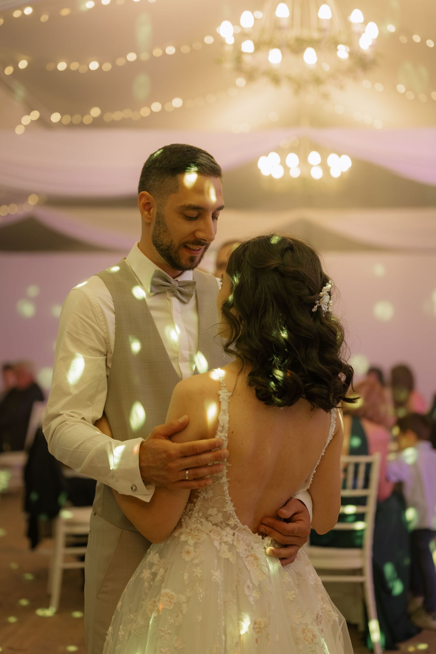 Bride and groom share first dance at elegant wedding reception with warm bokeh lights and chandelier backdrop.