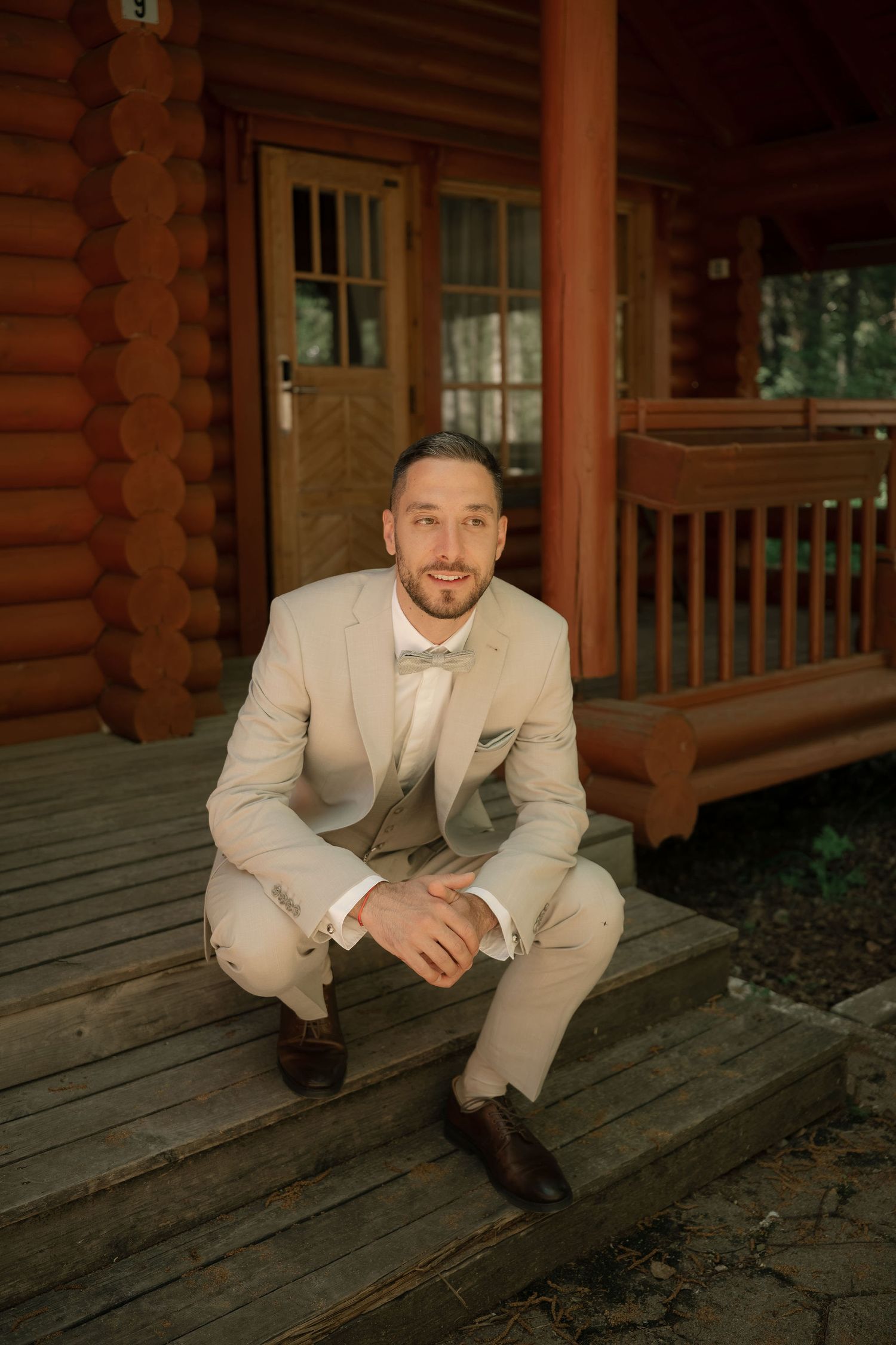 A well-dressed groom in a beige suit sits on wooden cabin porch steps, smiling confidently on his wedding day.