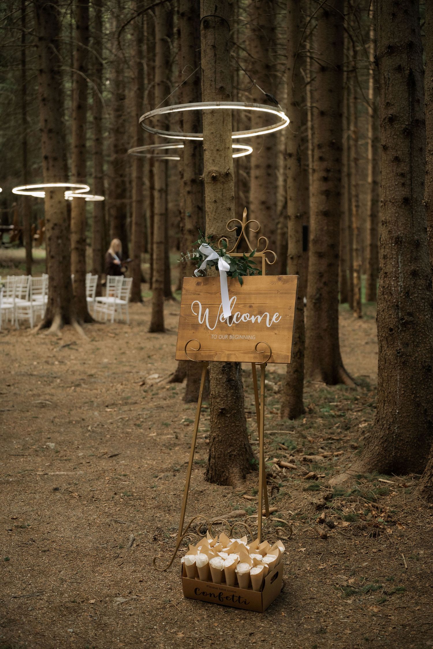 Rustic wooden welcome sign on easel in pine forest wedding venue with circular lights and white chairs.