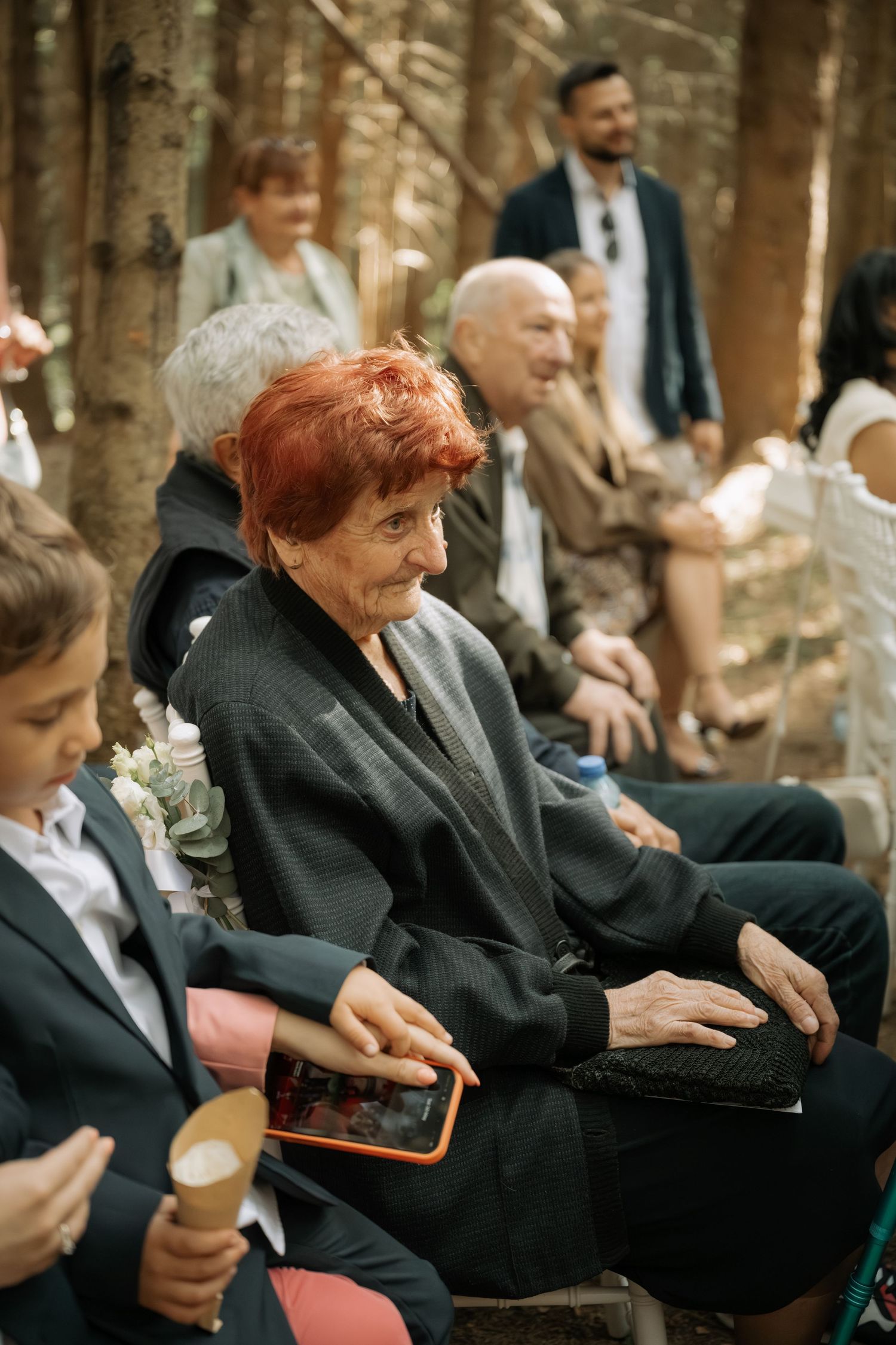 Elderly woman with red hair holds a tablet at an outdoor wedding ceremony in a forest setting.