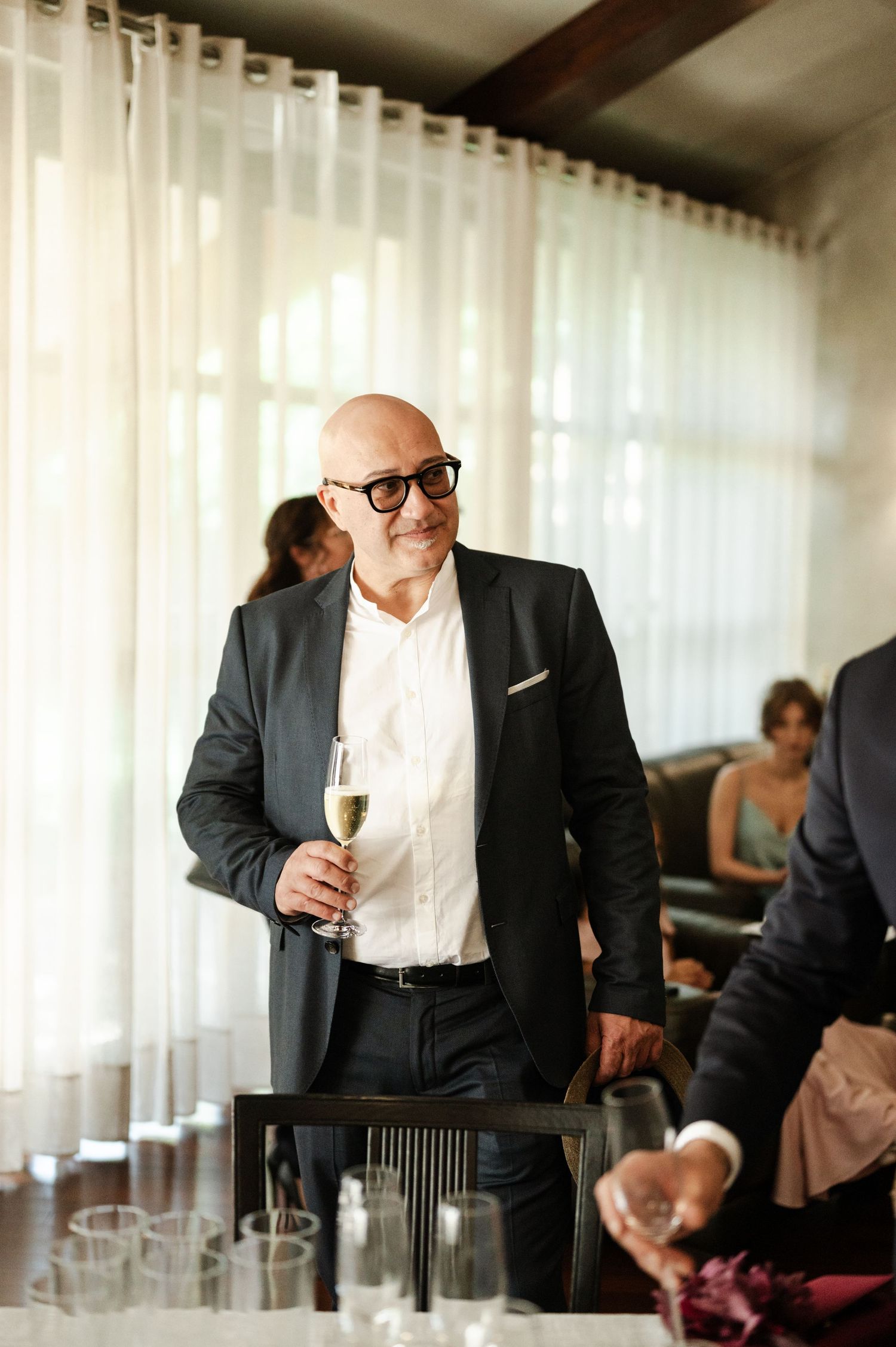 A guest in a dark suit smiles while holding a champagne glass at a wedding reception.