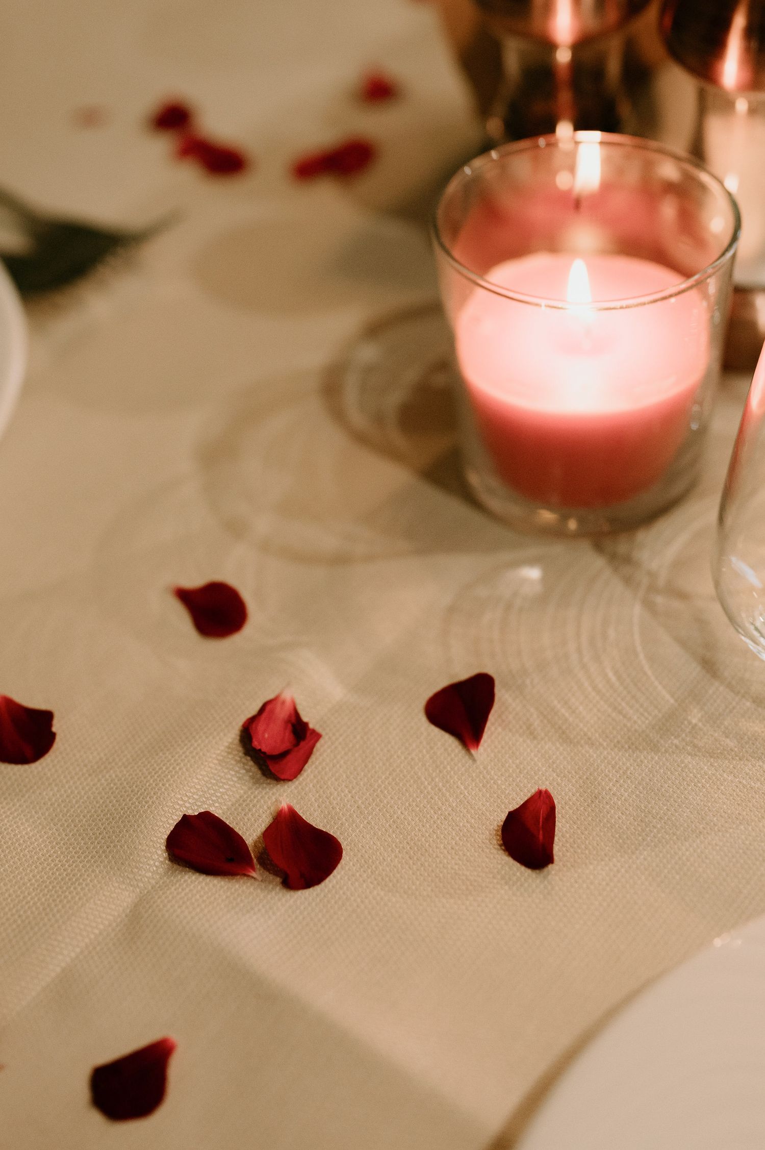 Pink candles and scattered red rose petals create a romantic ambiance on a white tablecloth.