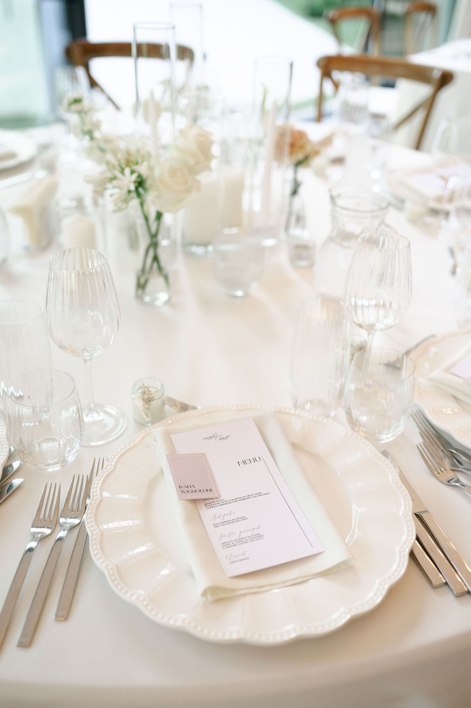 White place setting with scalloped plate, menu card, and delicate floral accents at wedding reception.