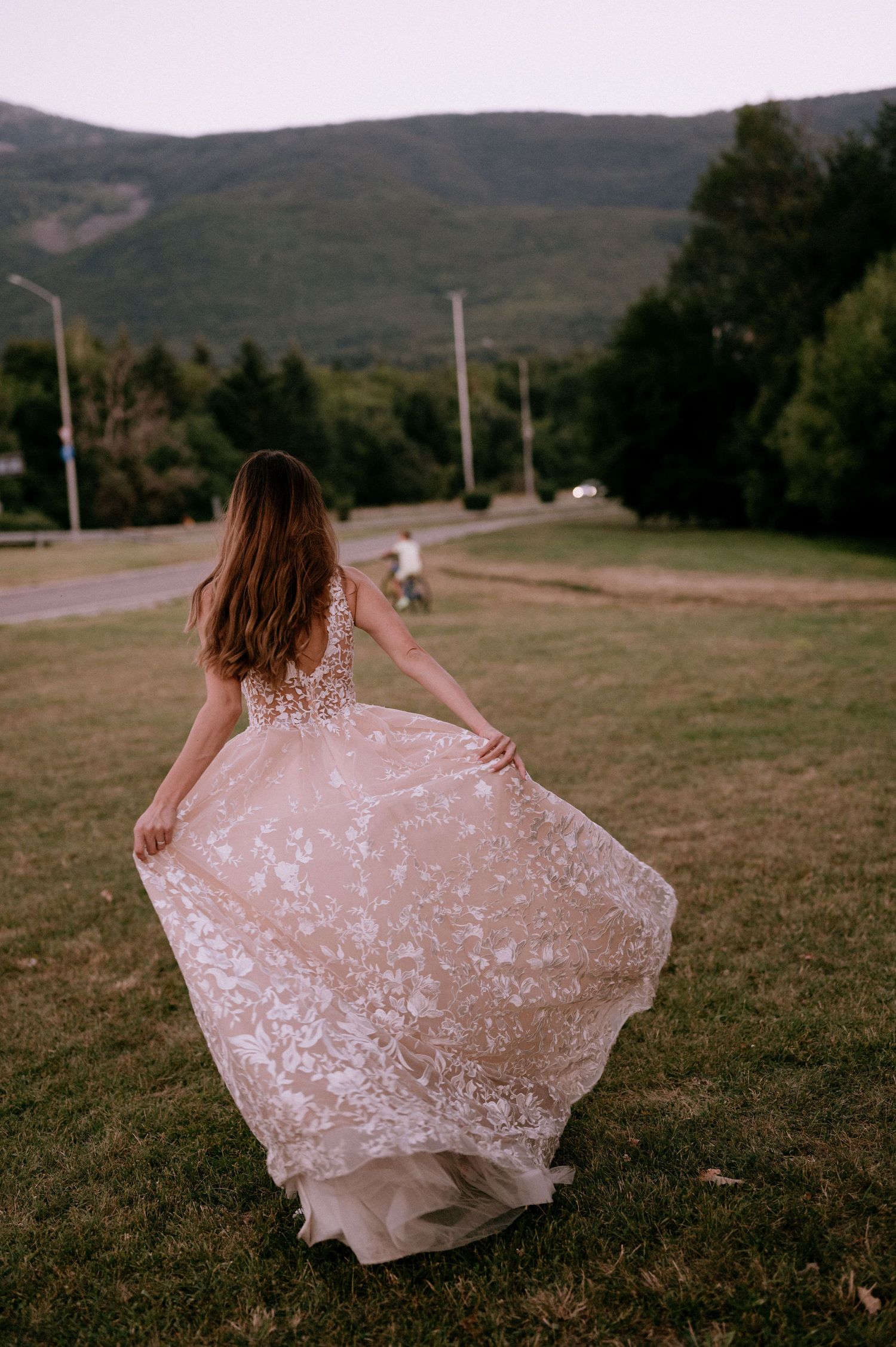Back view of bride in blush lace gown spinning in mountain field at sunset.