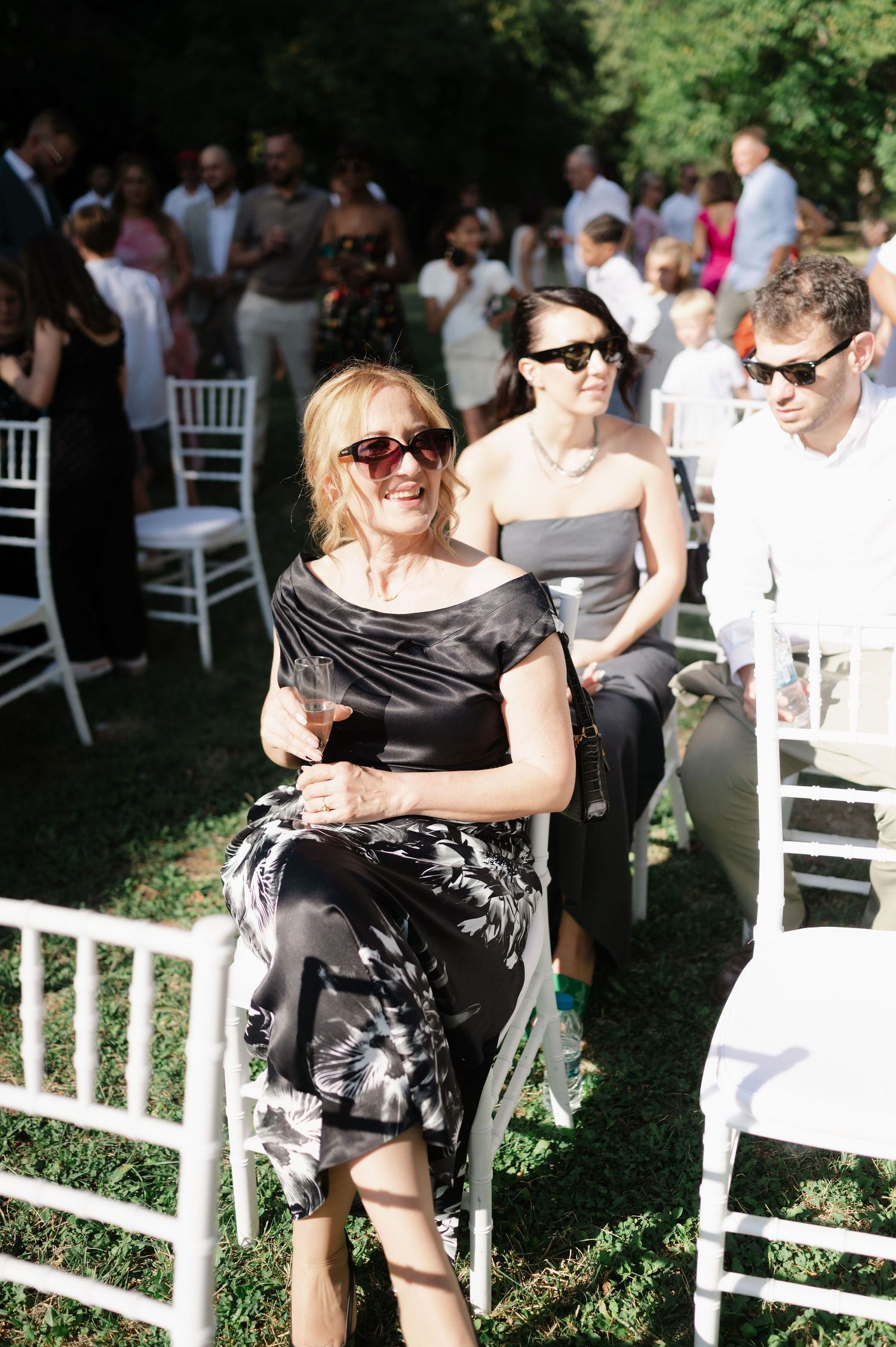 Wedding guests in sunglasses gather on white chairs during an outdoor summer ceremony.