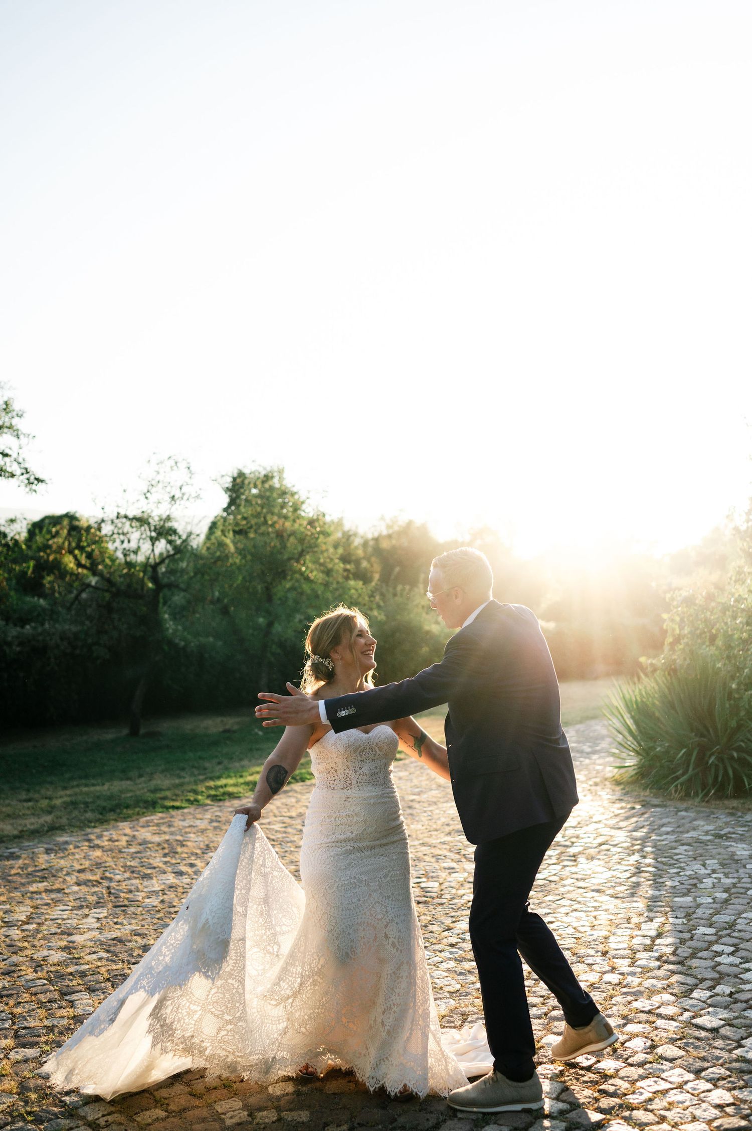 Playful wedding couple spinning and dancing together at sunset on stone pathway.