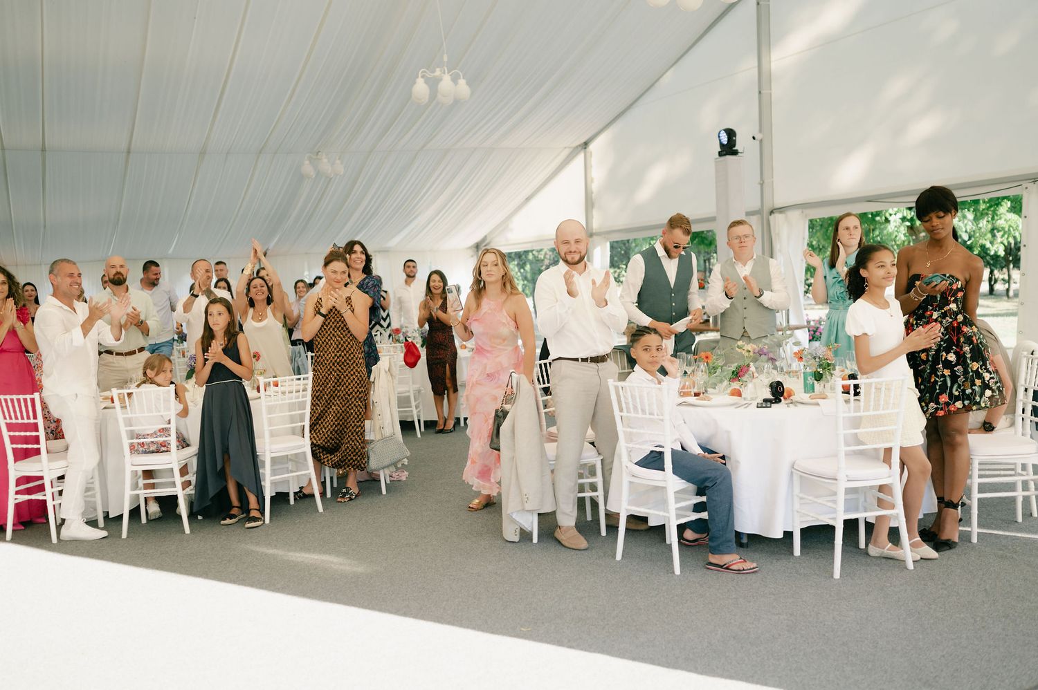 Wedding guests seated at round tables in white tent for reception celebration.