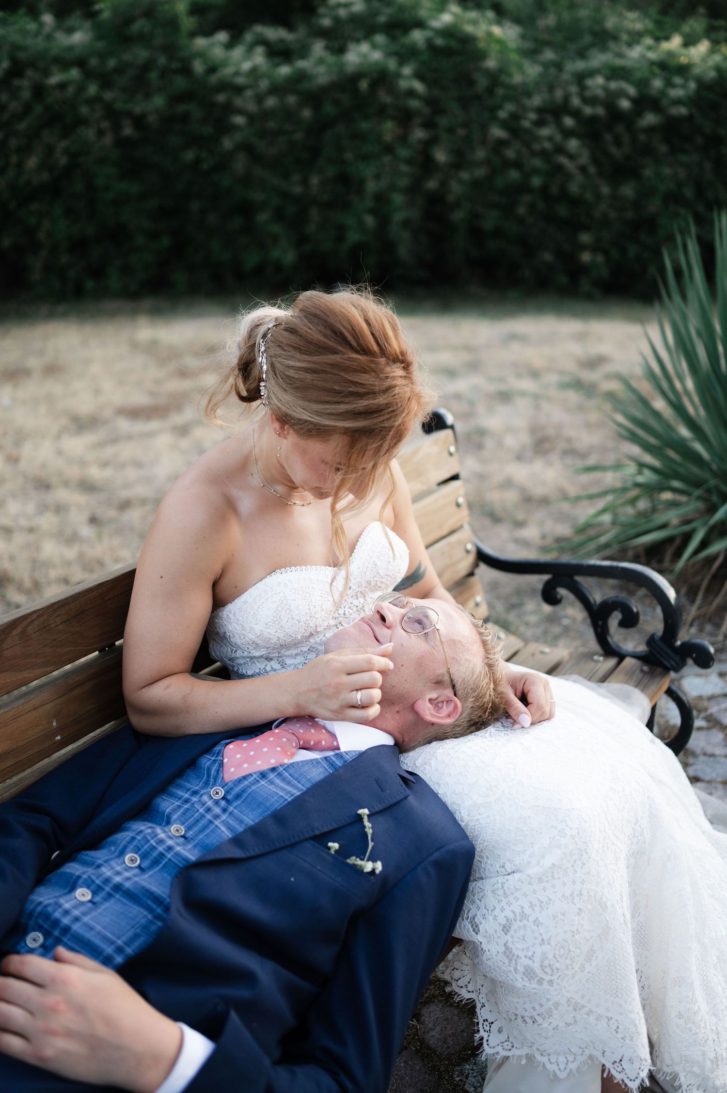 Newlyweds share a tender moment while resting on a wooden bench in a garden setting.