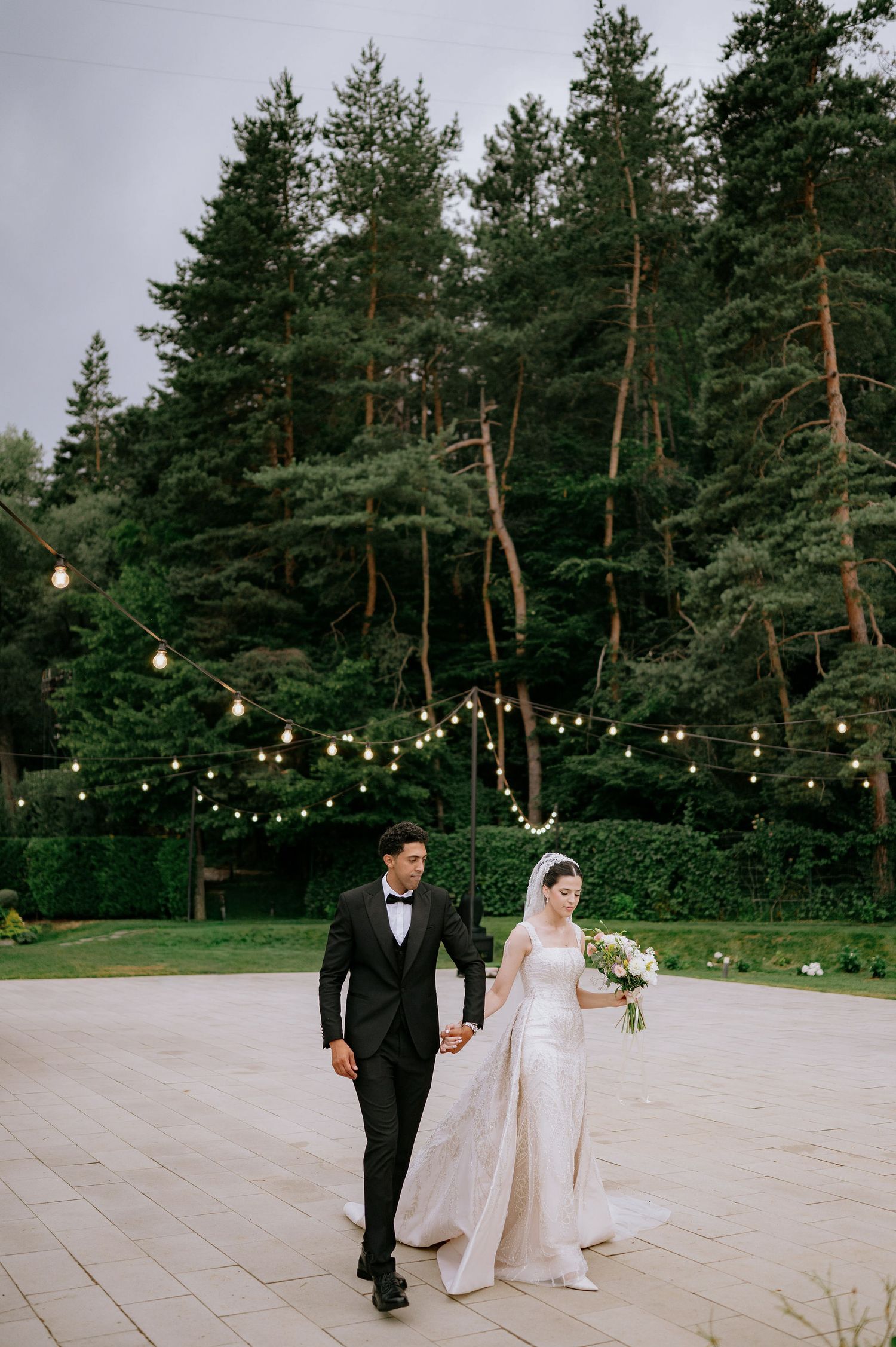 A couple in wedding attire walks along a path with string lights and evergreen trees in the background.