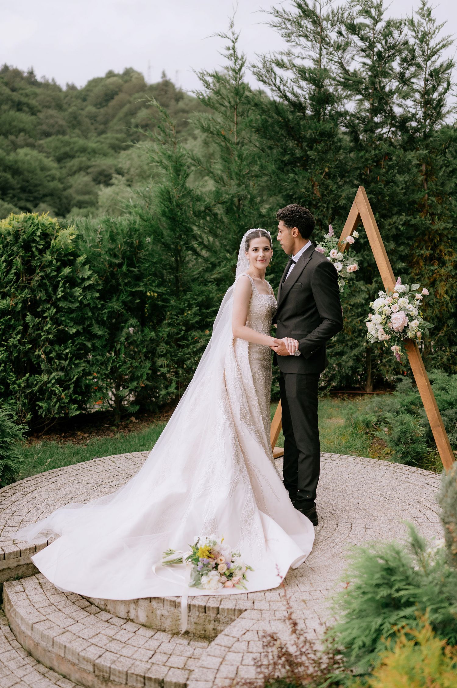 Couple exchanging vows on stone patio surrounded by evergreen trees and wooden ceremony arch.